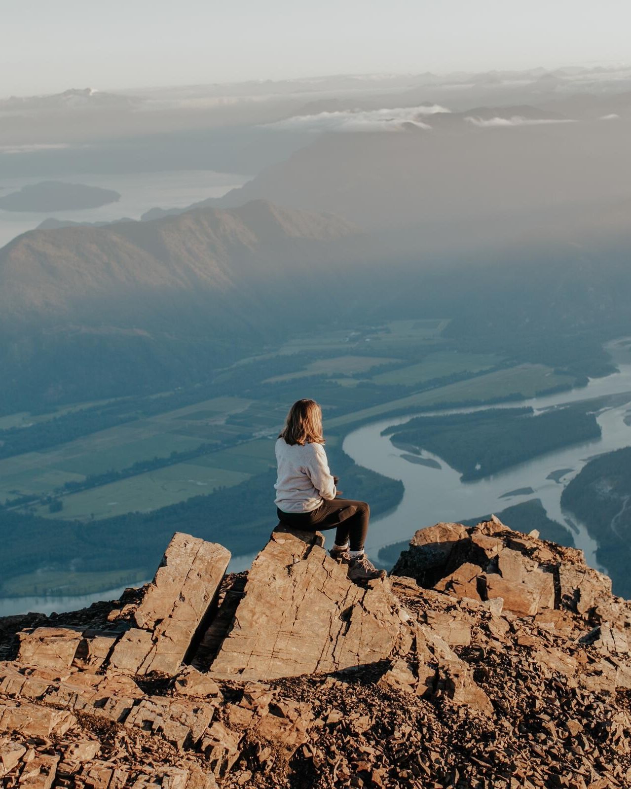 A hiker sitting on a rocky summit above a winding river valley.