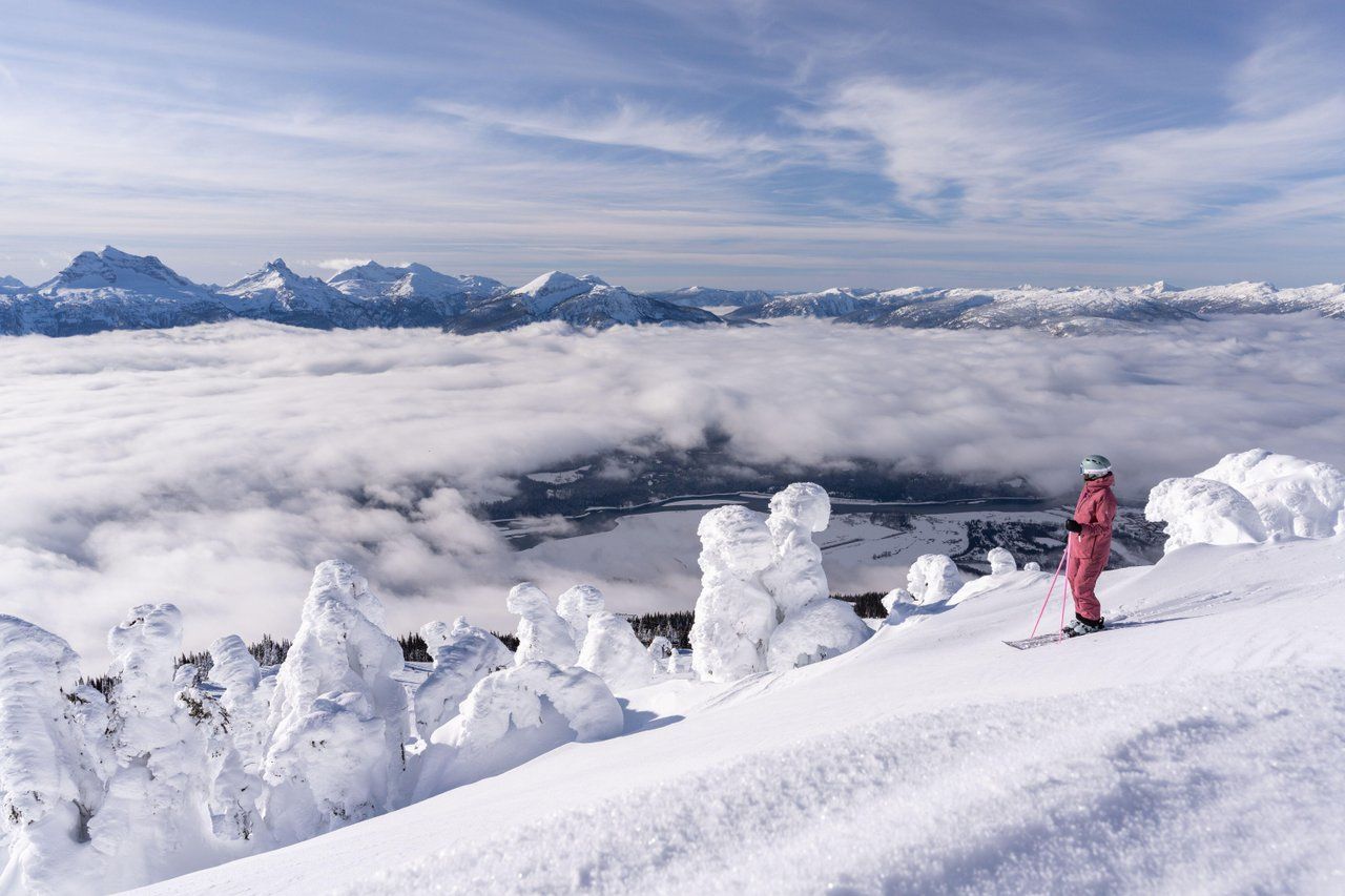 Skier overlooking snow-covered mountains and clouds from an alpine ridge.