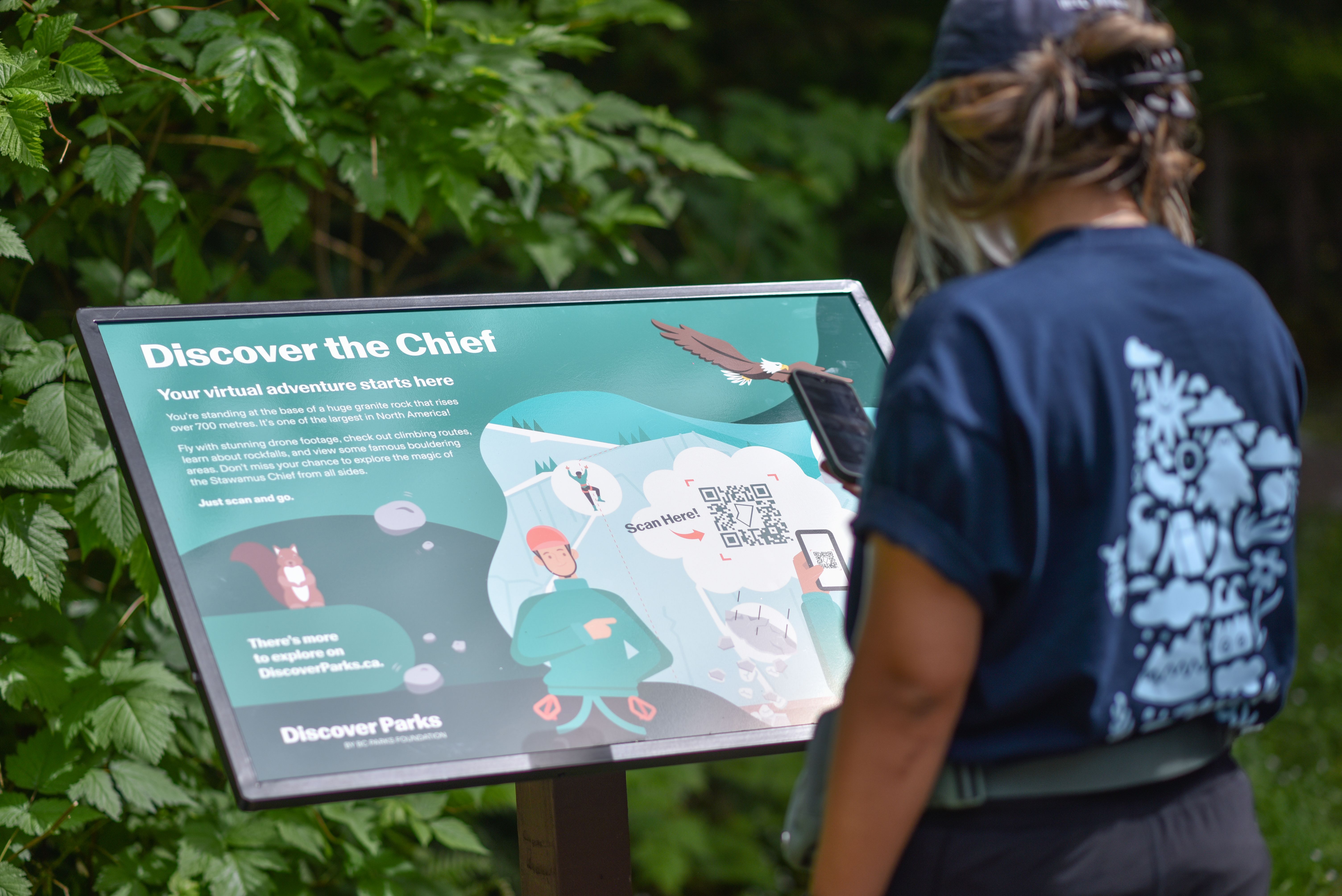 Woman uses phone to scan a QR code on a Discover Trail sign saying "Discover the Chief"