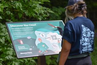 Woman uses phone to scan a QR code on a Discover Trail sign saying "Discover the Chief"