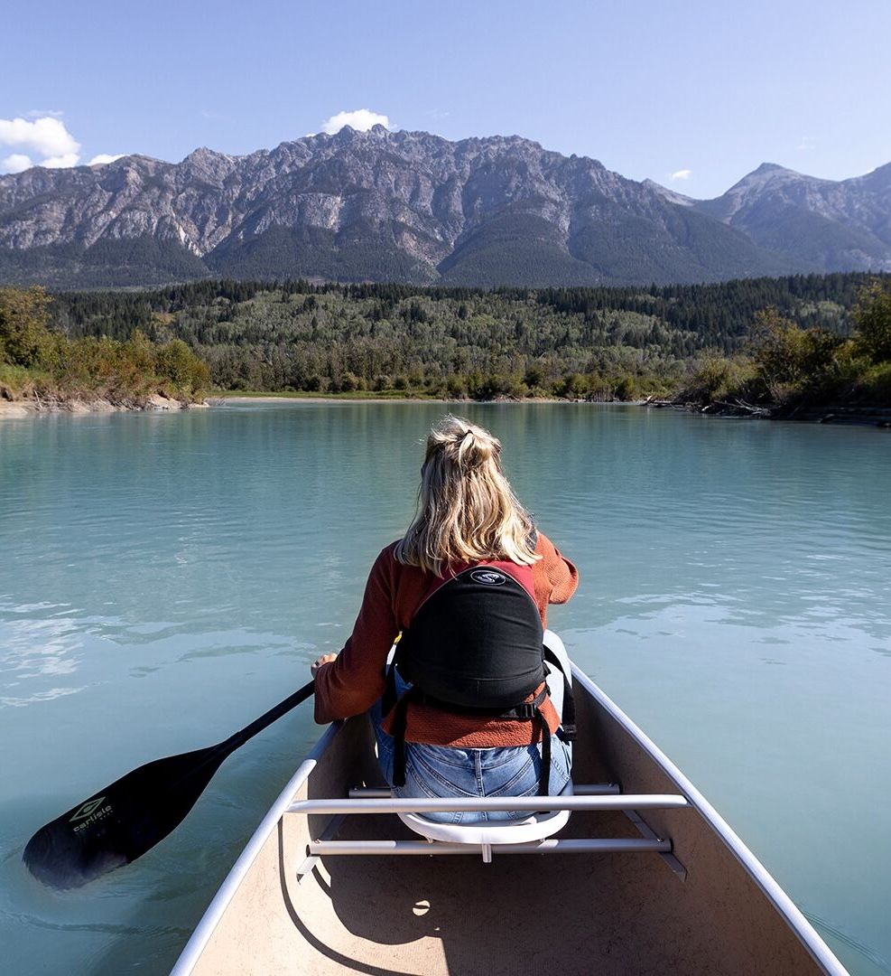 A woman paddling a canoe on a turquoise river toward rugged mountains.