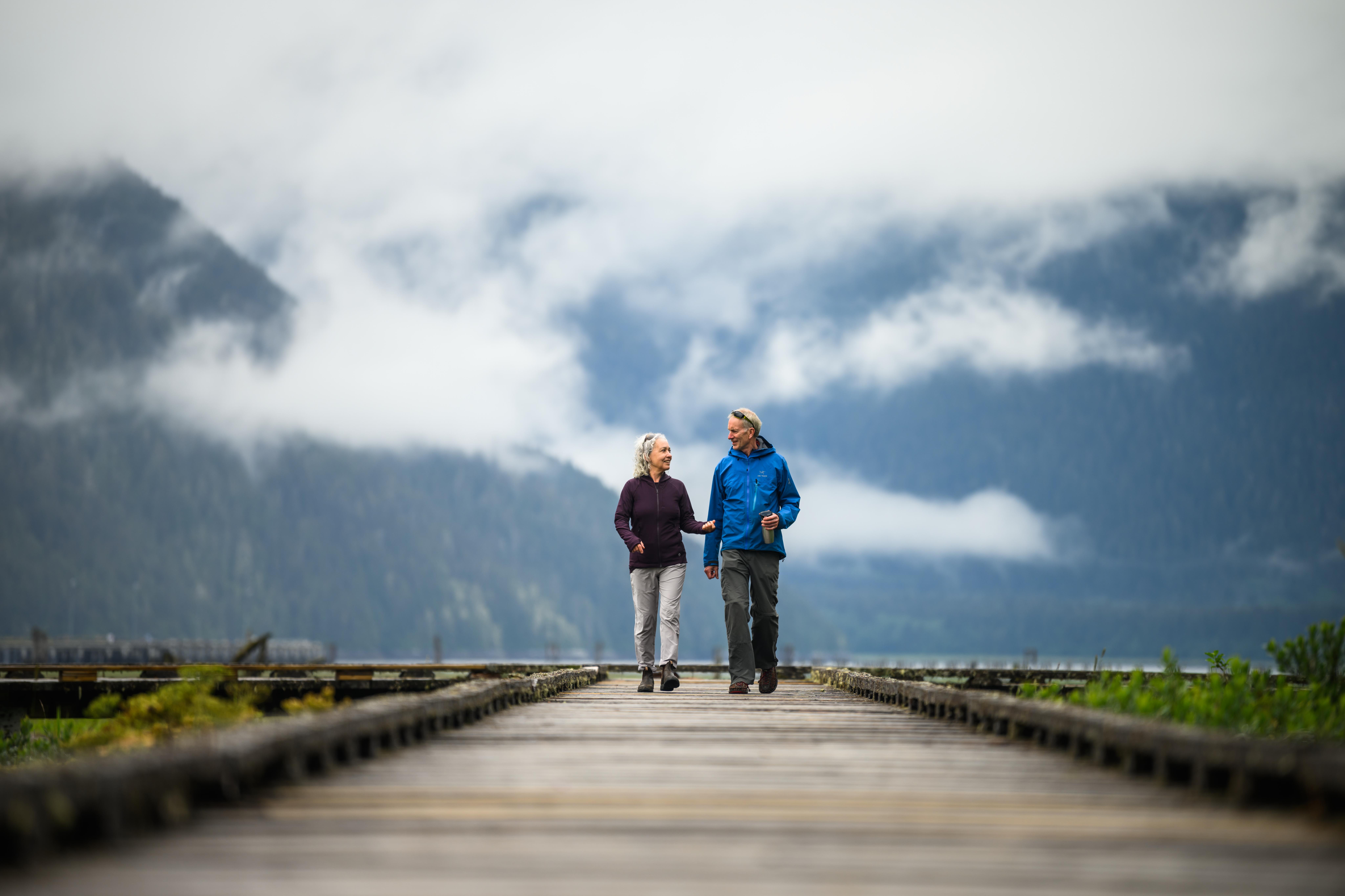 A couple stroll across a bridge with mountains in a cloudy setting in the surroundings