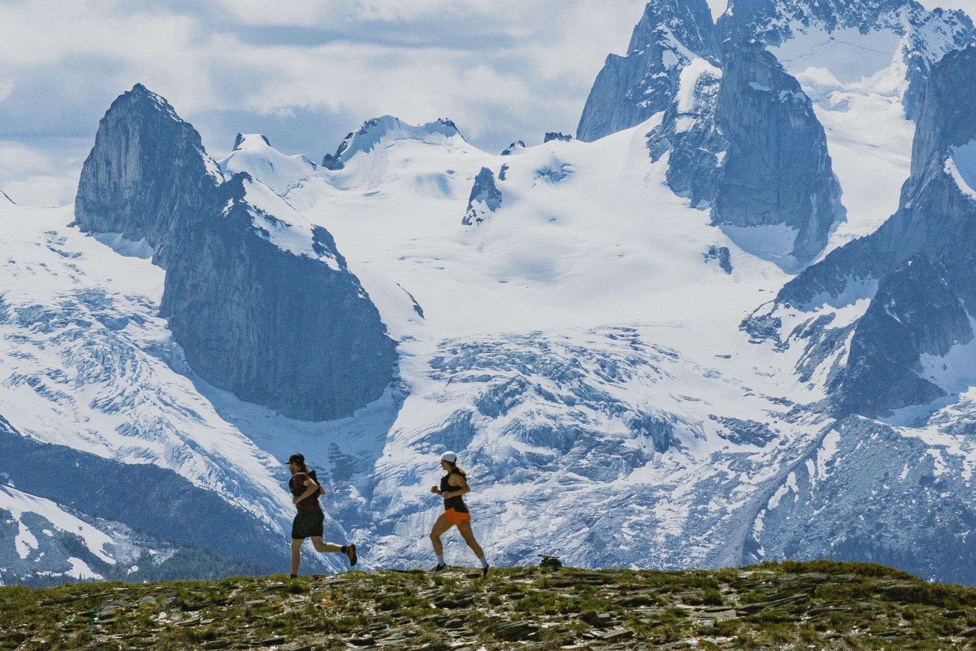 Two trail runners crossing an alpine ridge with towering snow-covered mountains and glaciers in the background.