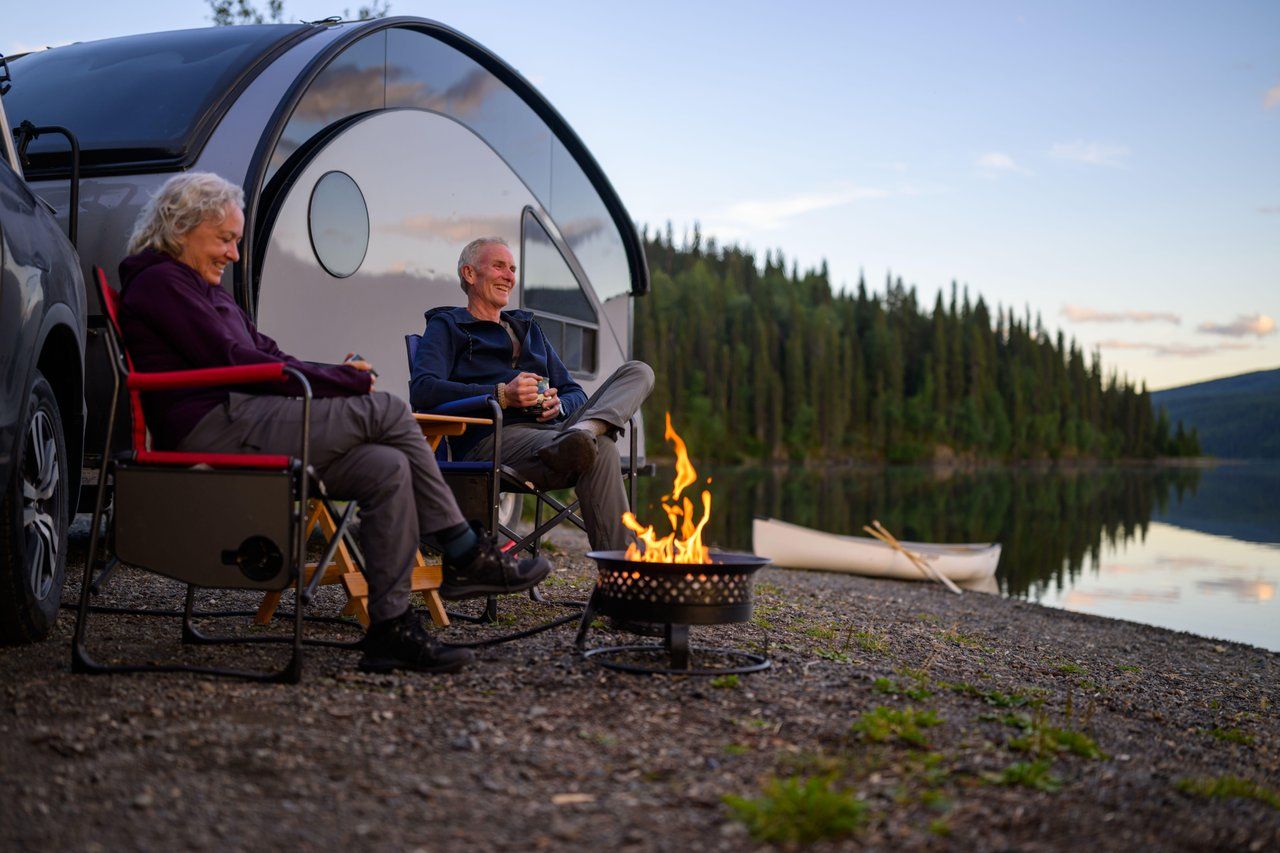 Two people relaxing beside a lakeside campsite with a campfire and canoe nearby.