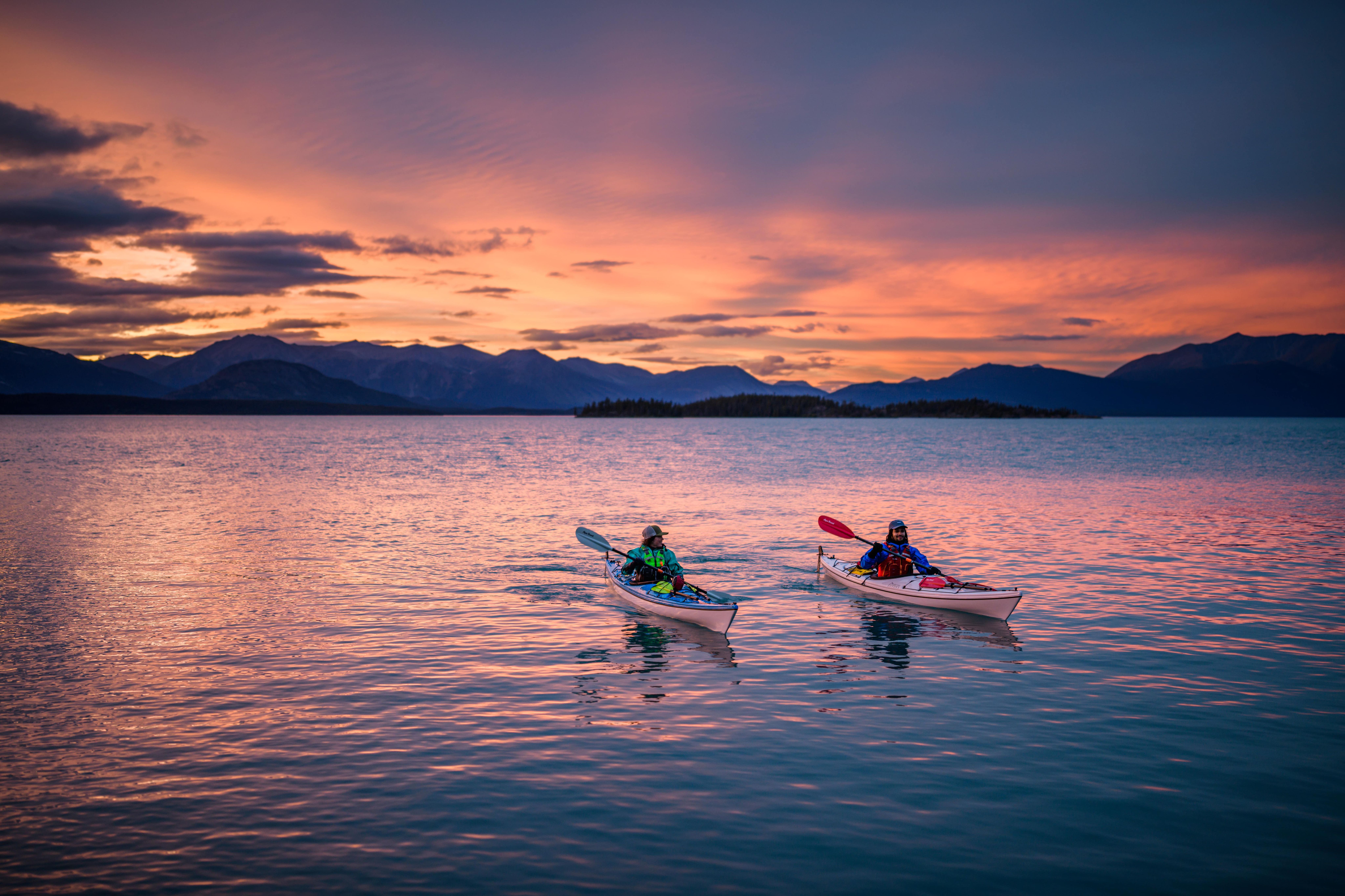 Two people kayak underneath a pink and orange setting sun