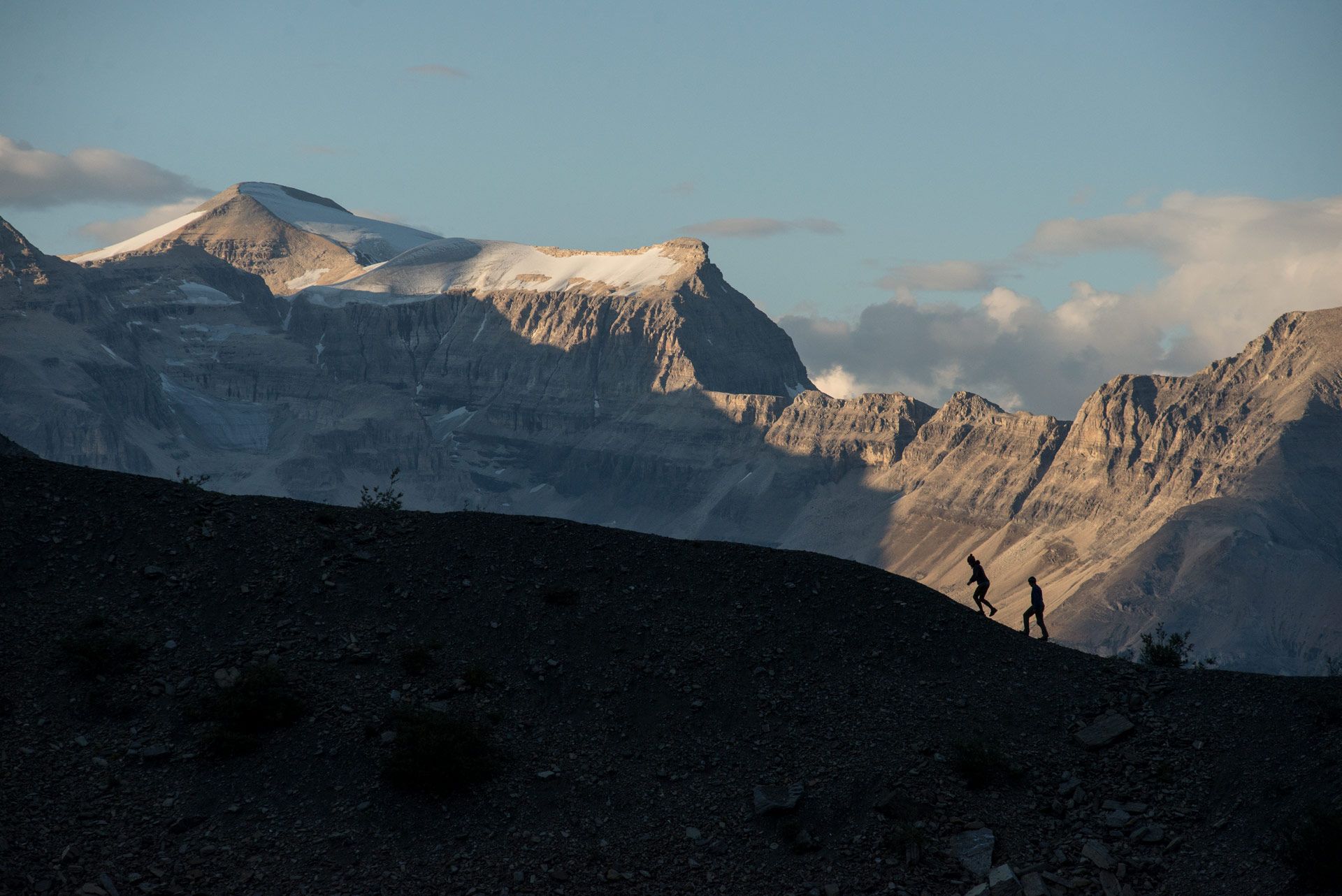 Two people traverse a mountain, with large snowcapped mountains in the background