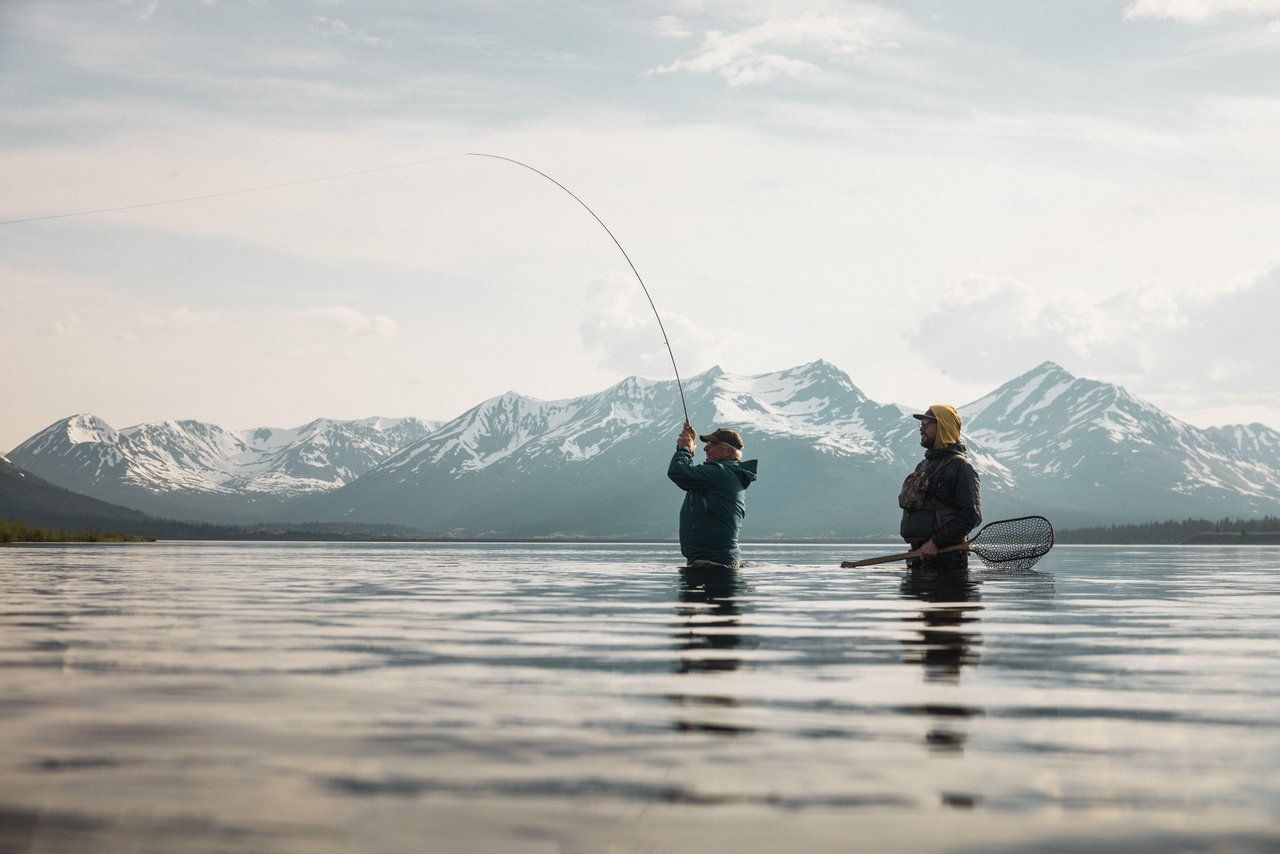 Two people fly fishing in a mountain lake with snow-covered peaks in the background.