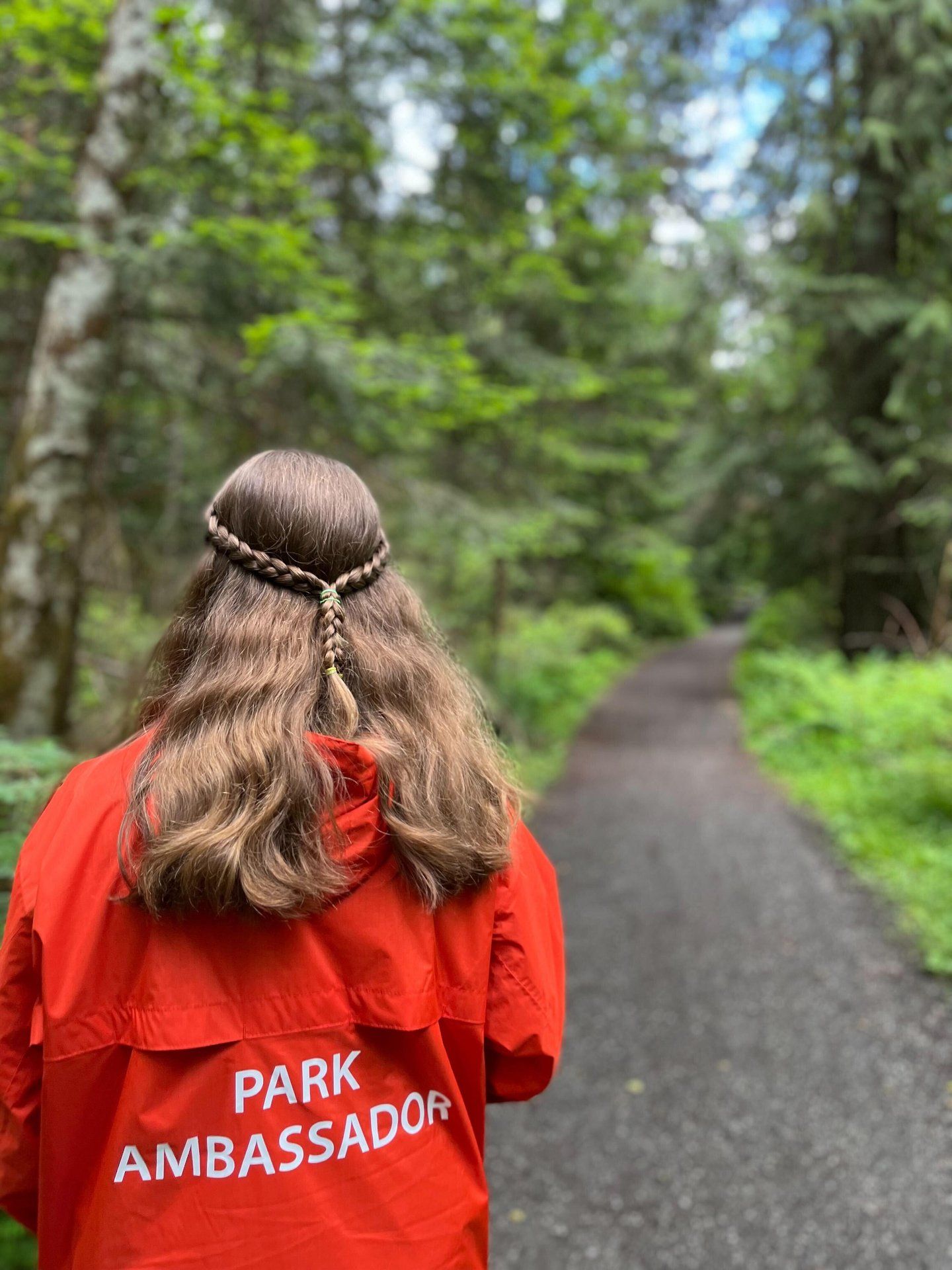 A Park Ambassador in a red jacket on a gravel trail through a green forest.