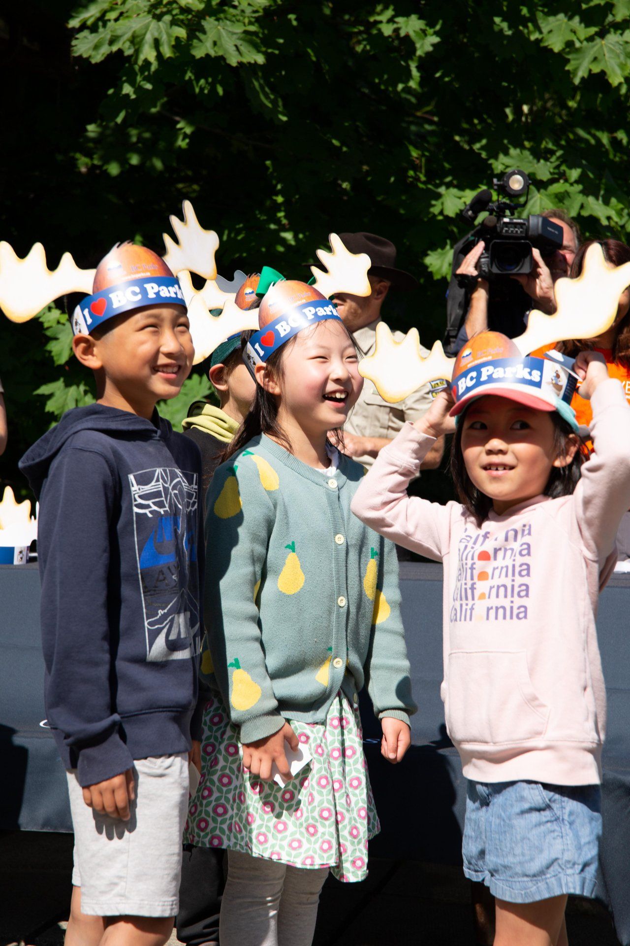     Three children wearing BC Parks moose antler hats smile and play together at an outdoor event