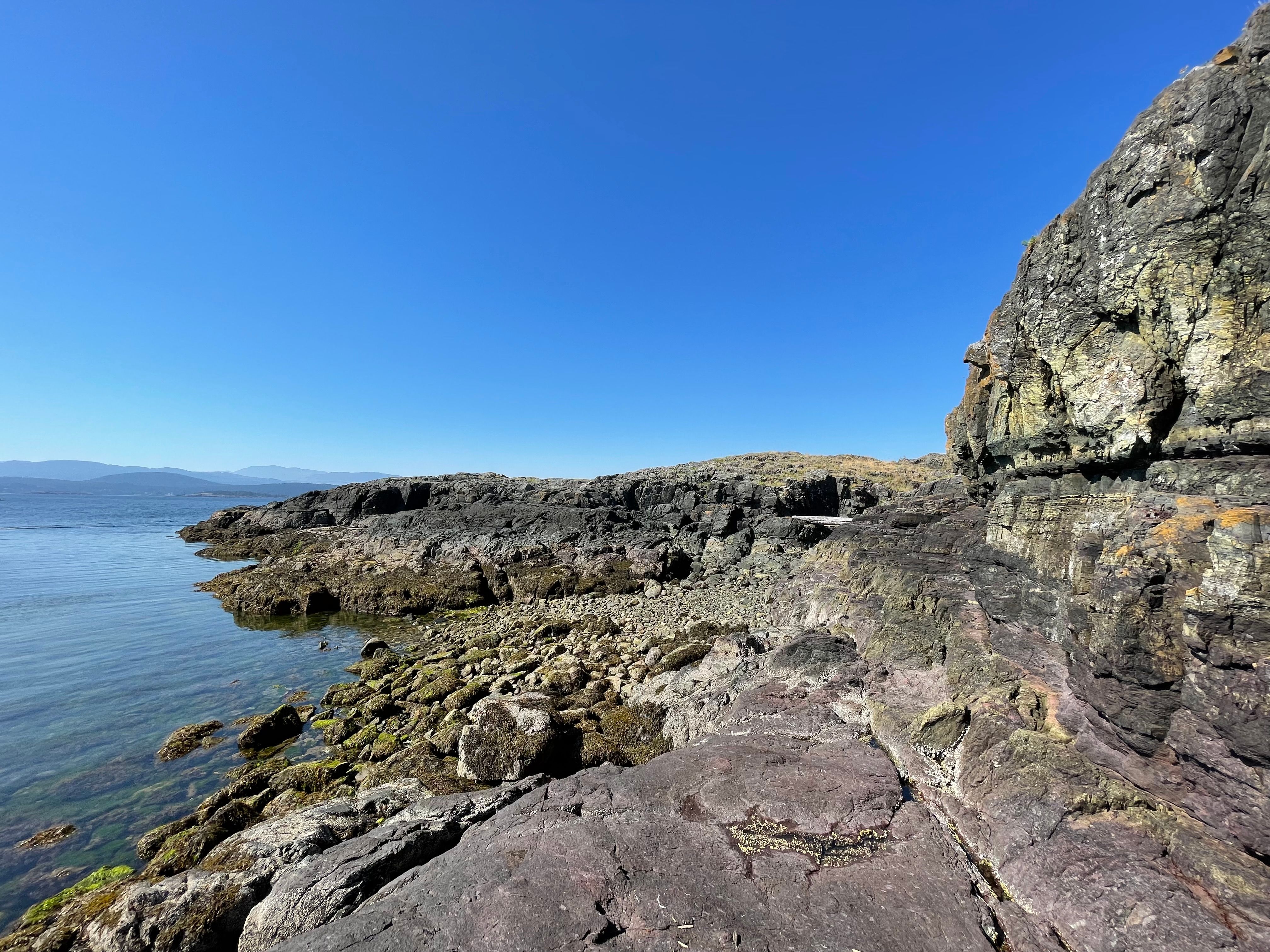 A wide lense of a rocky shoreline with shrubs, clouds and blue skies in shot