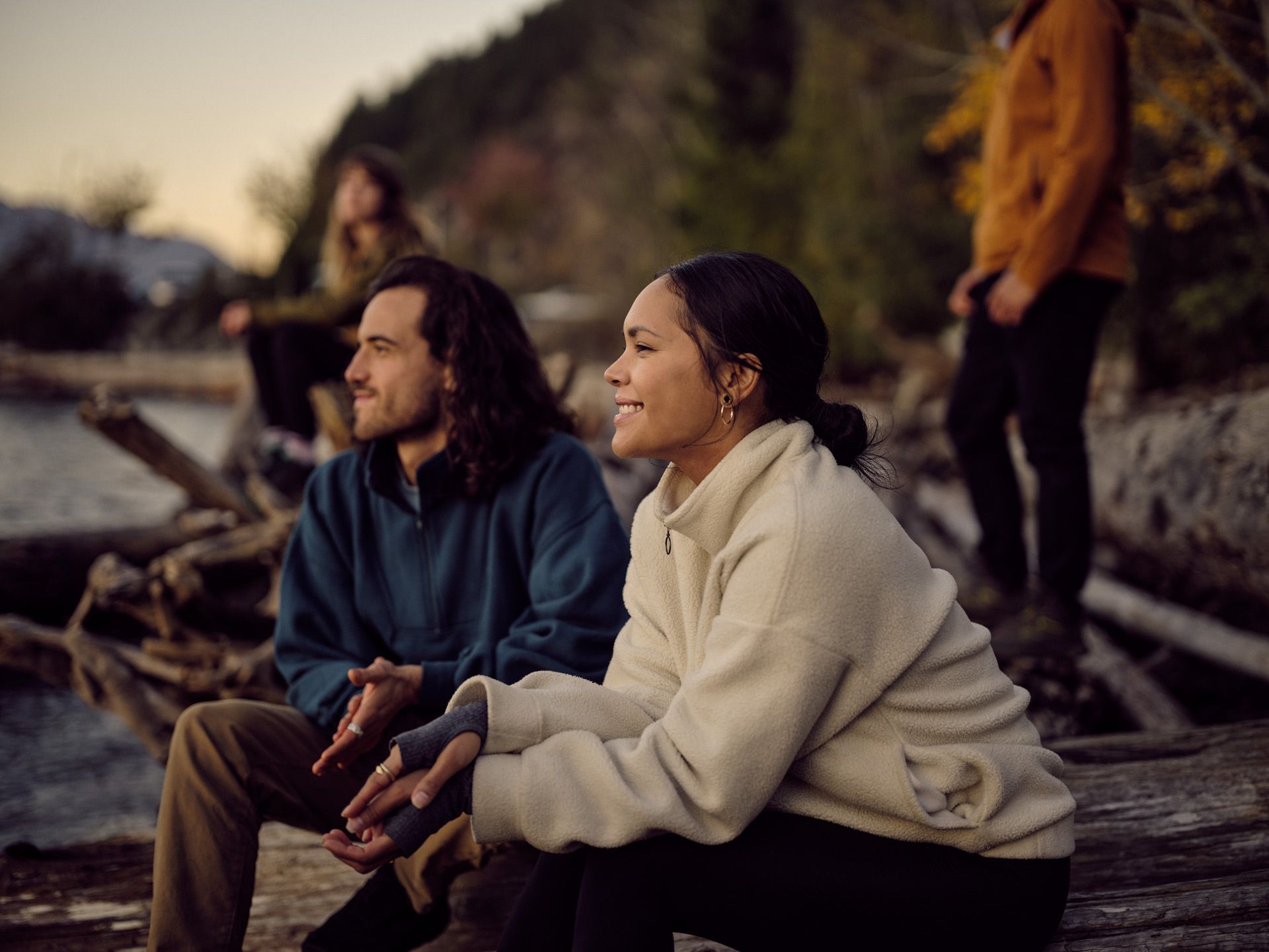 Two smiling people sit down in a beach setting with more people in the background