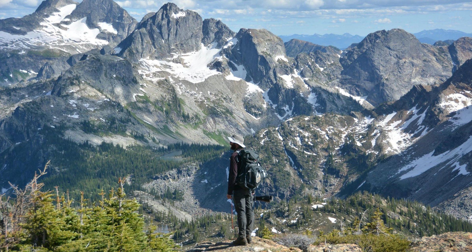 Hiker standing on a mountain ridge overlooking snow-capped mountains and forest.