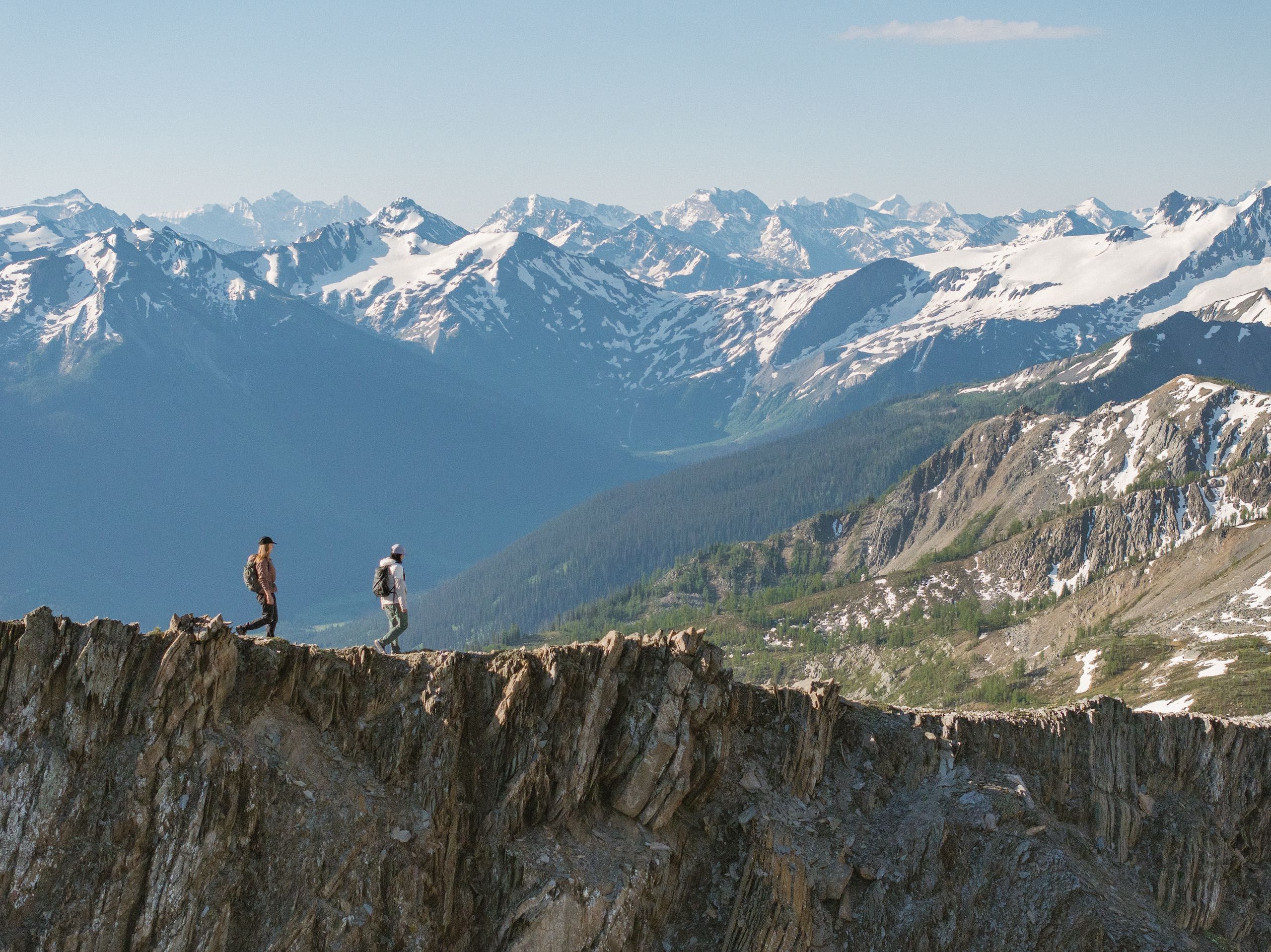 Two people traverse a rock face with snow-capped mountains in the background
