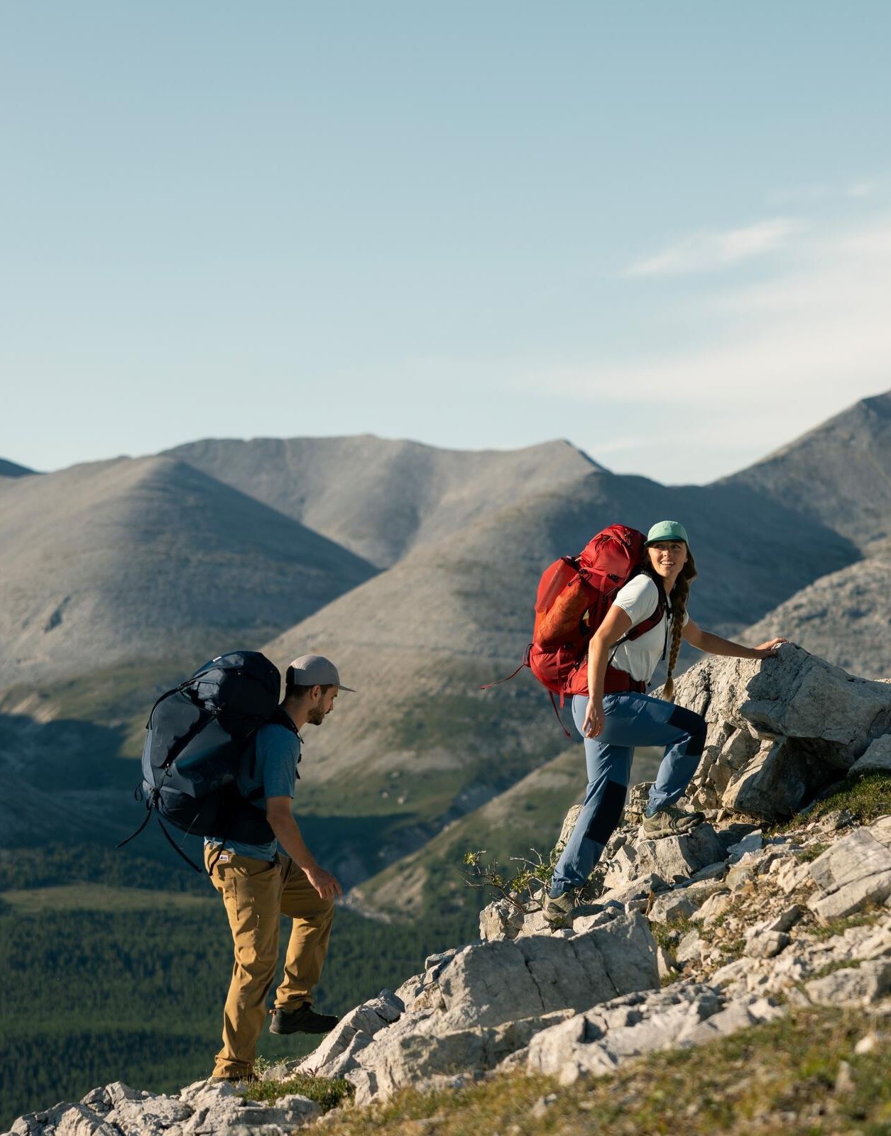 Two backpackers scrambling up a rocky ridge with bare grey mountains behind them.