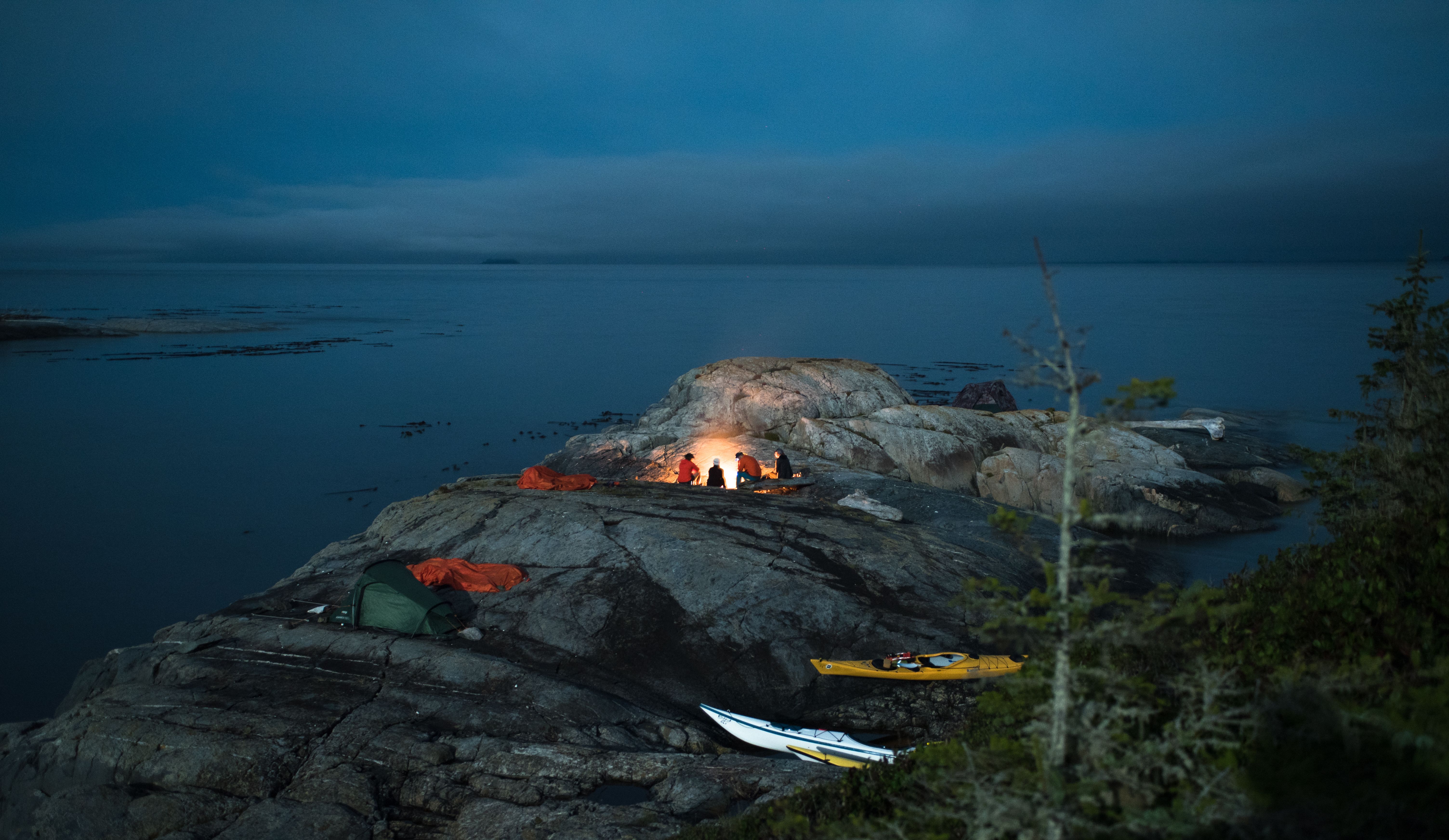 A group of kayakers gather around a fire pit on a rocky island