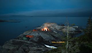 A group of kayakers gather around a fire pit on a rocky island