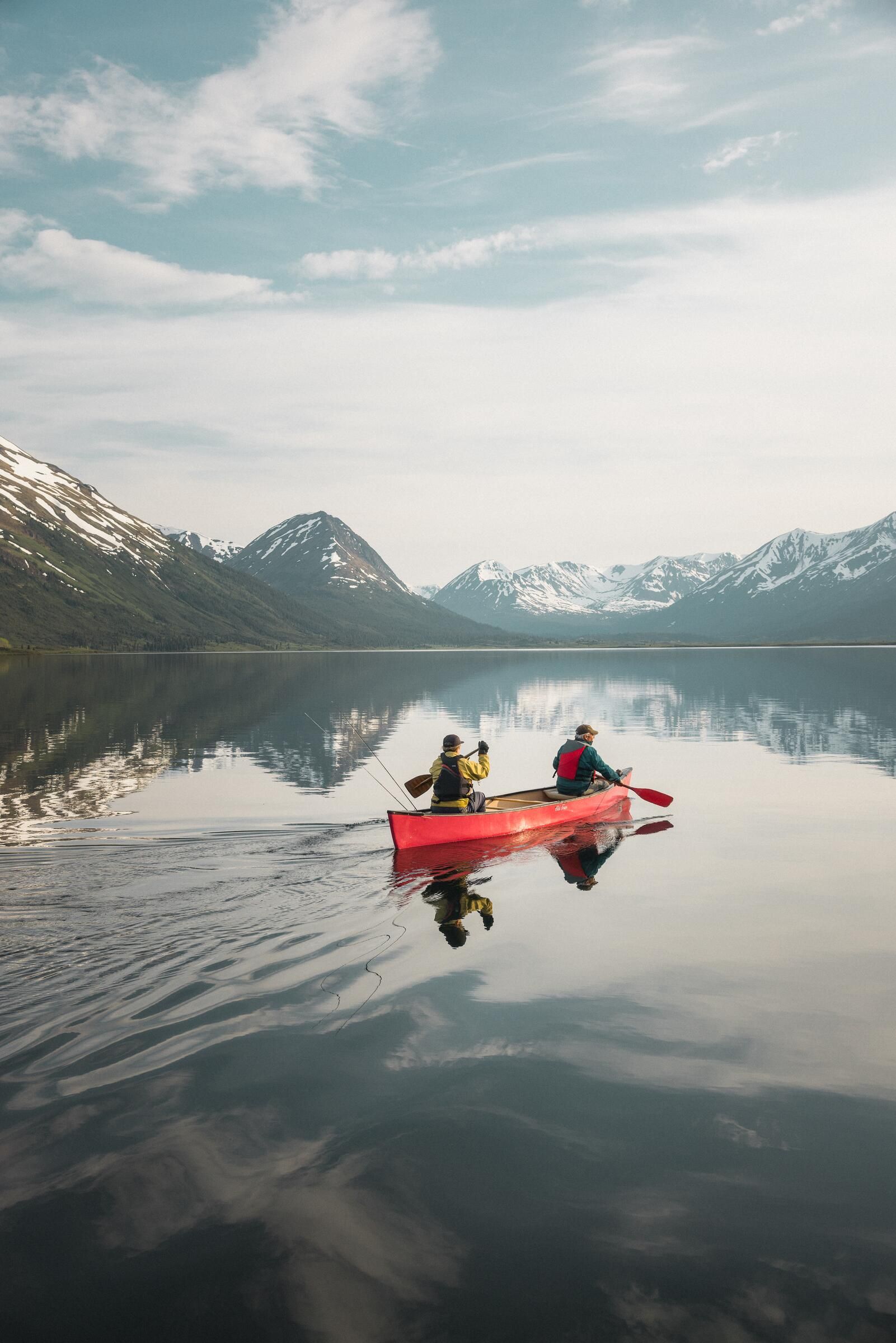 Two people in a red canoe on a still mountain lake, with snowy peaks reflected in the water.