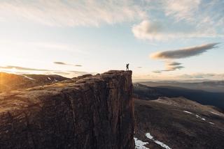 A person stands atop a large cliff, overlooking a deep valley