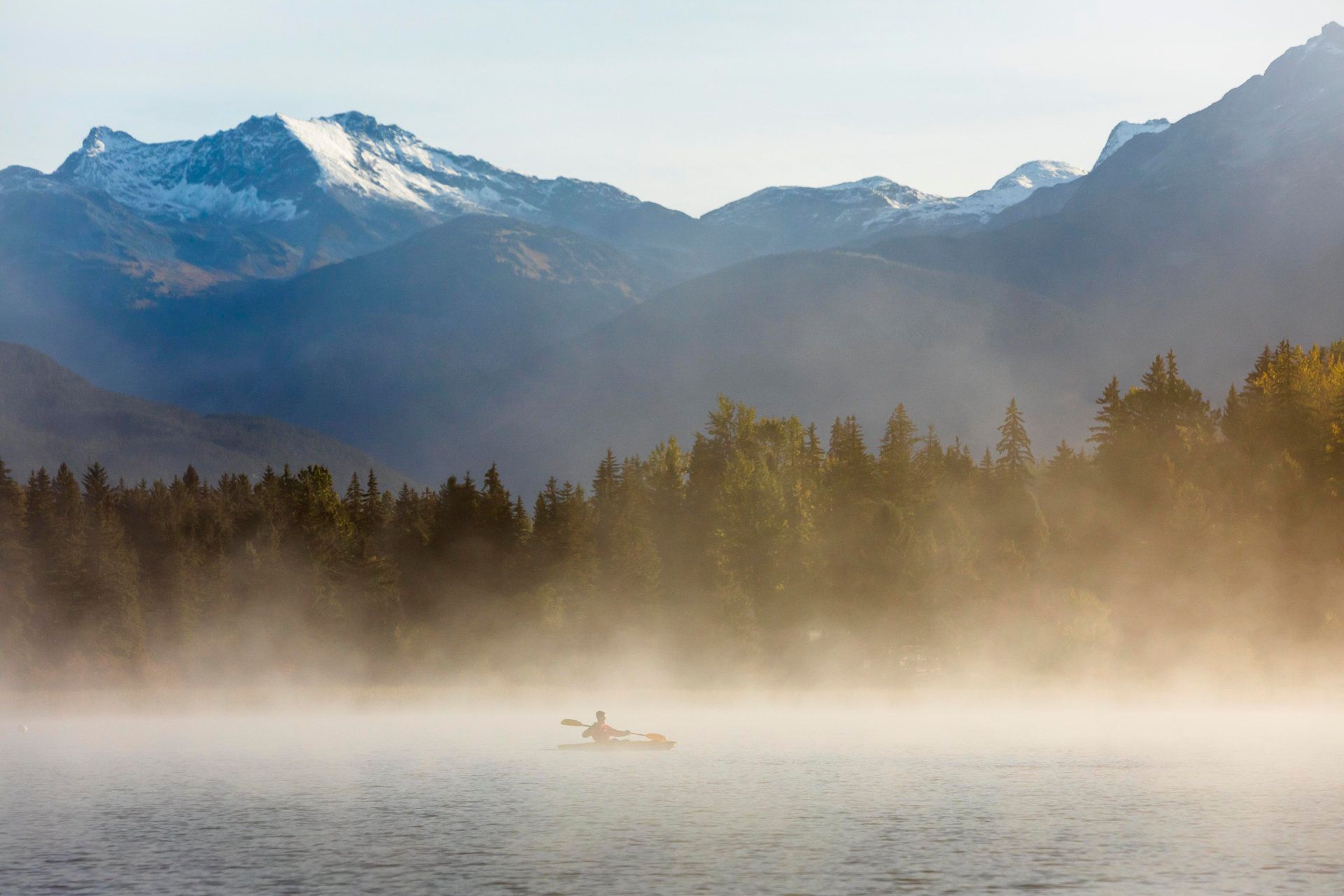 Kayaker paddling across a misty lake with snow-capped mountains in the background.