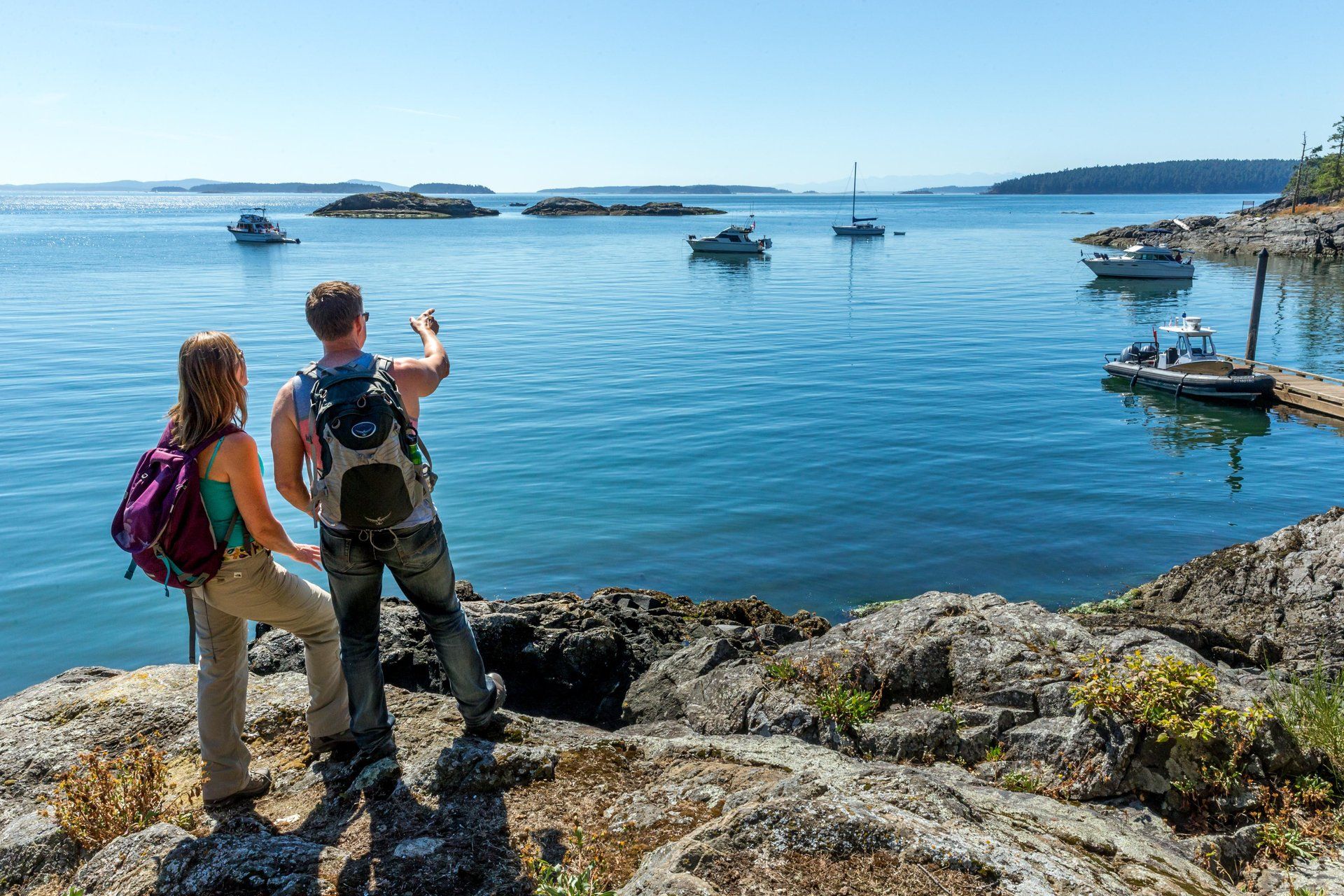 Two hikers with backpacks stand on rocky shoreline.