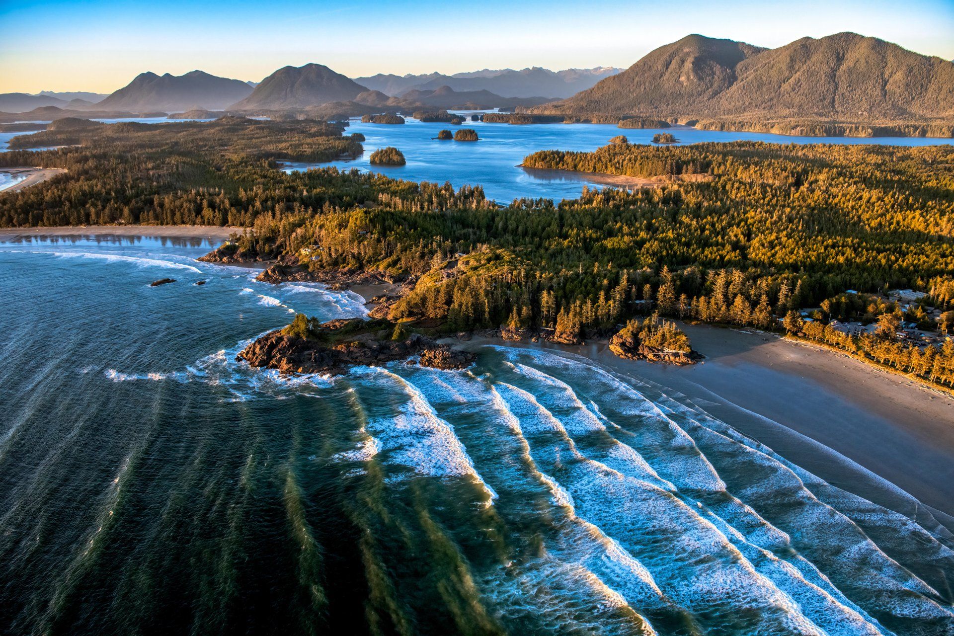 Aerial view of a sandy coastline, forested islands, and mountains beside the ocean.