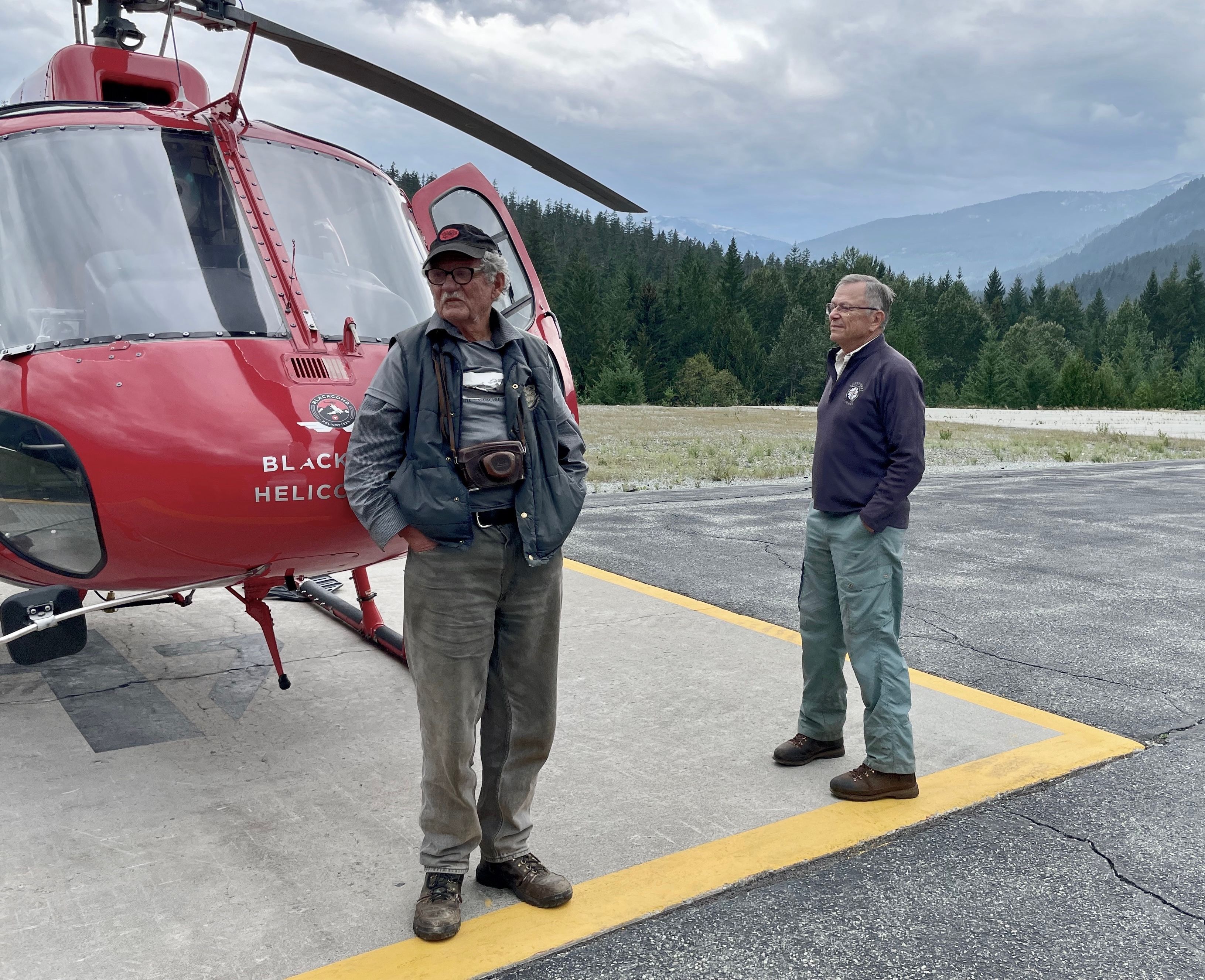 Two people standing beside a red helicopter in a mountain valley clearing.