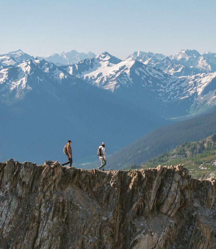 Two people traverse a rock face with snow-capped mountains in the background