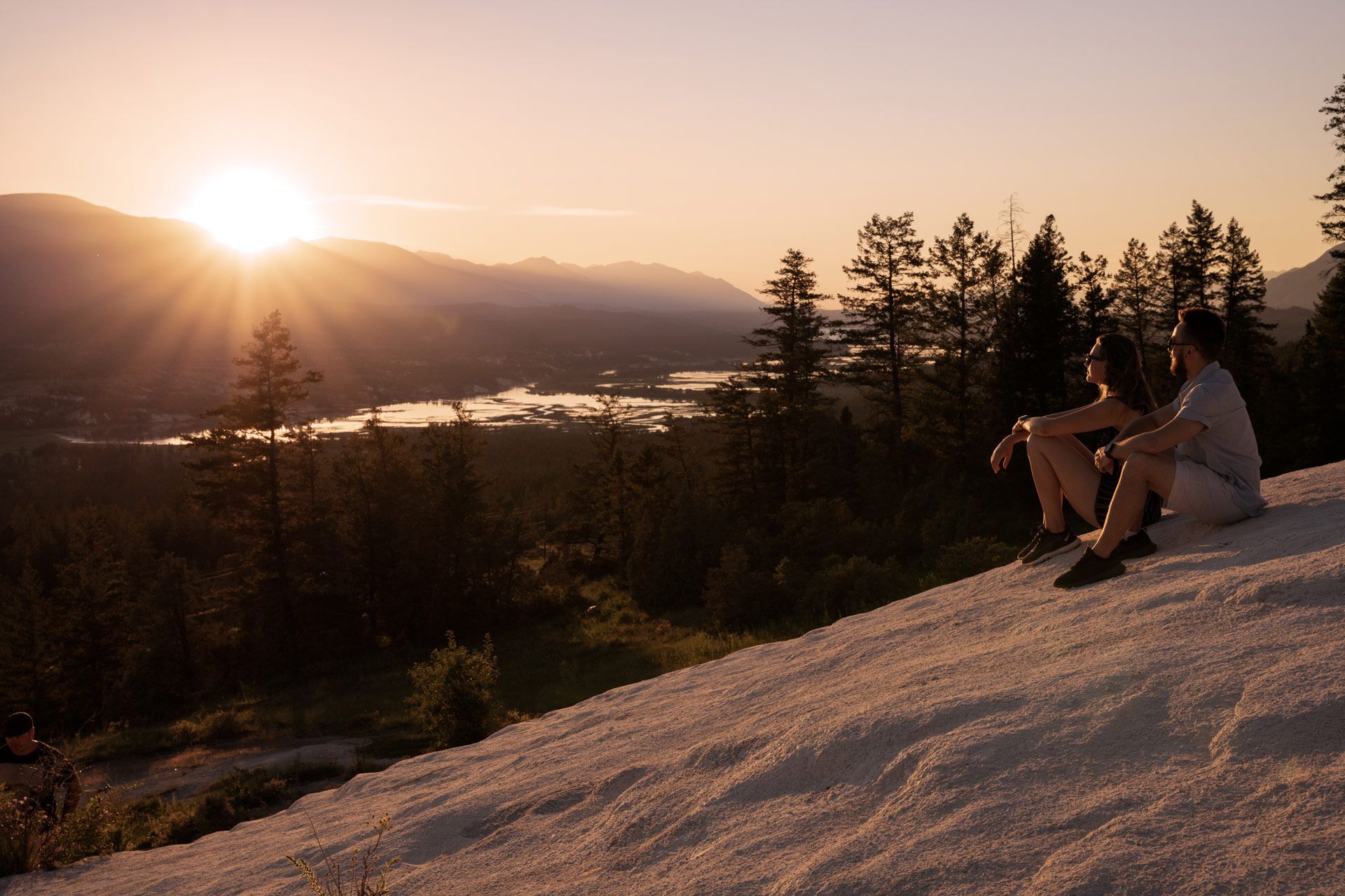 Two people sit on a mountainside and overlook a valley with a sun setting in the distance