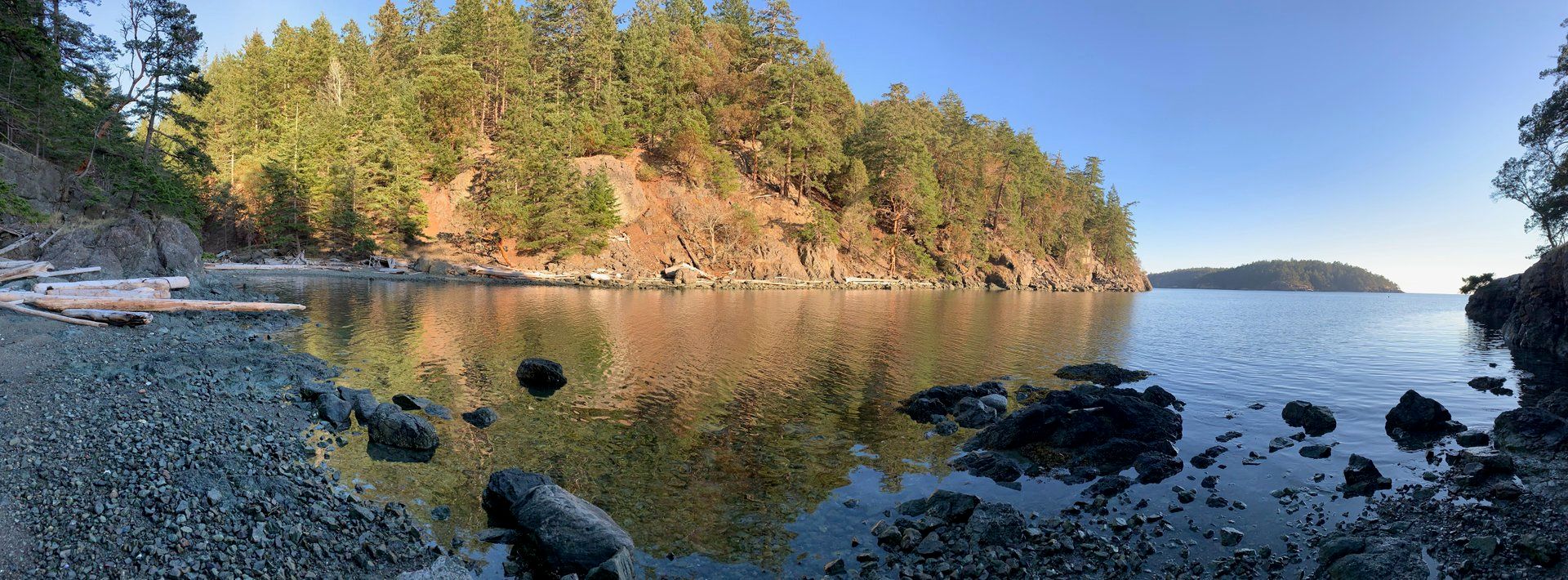 A calm, rocky cove with a sunlit forested bluff reflected in the water.