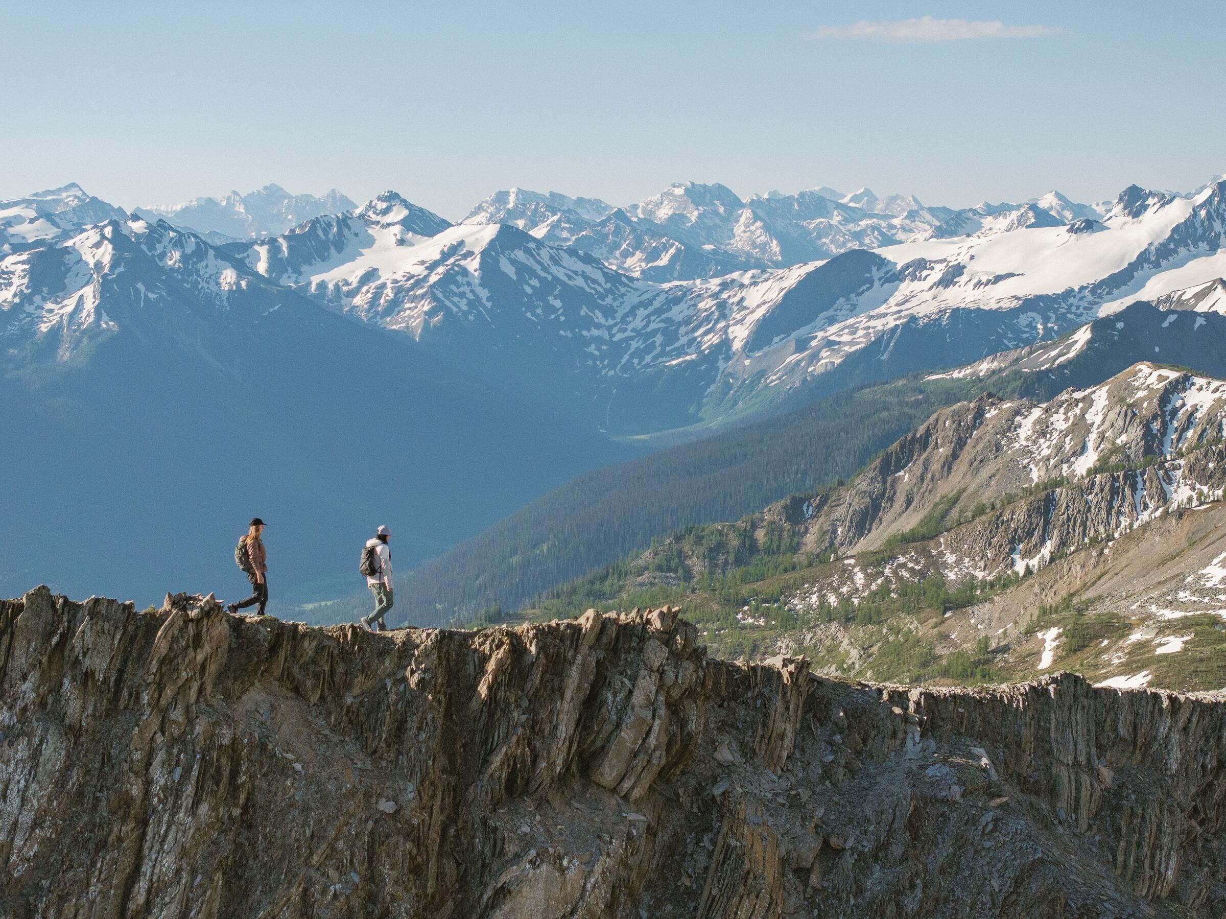 Two hikers walking along a rocky mountain ridge with snow-capped peaks in the distance.