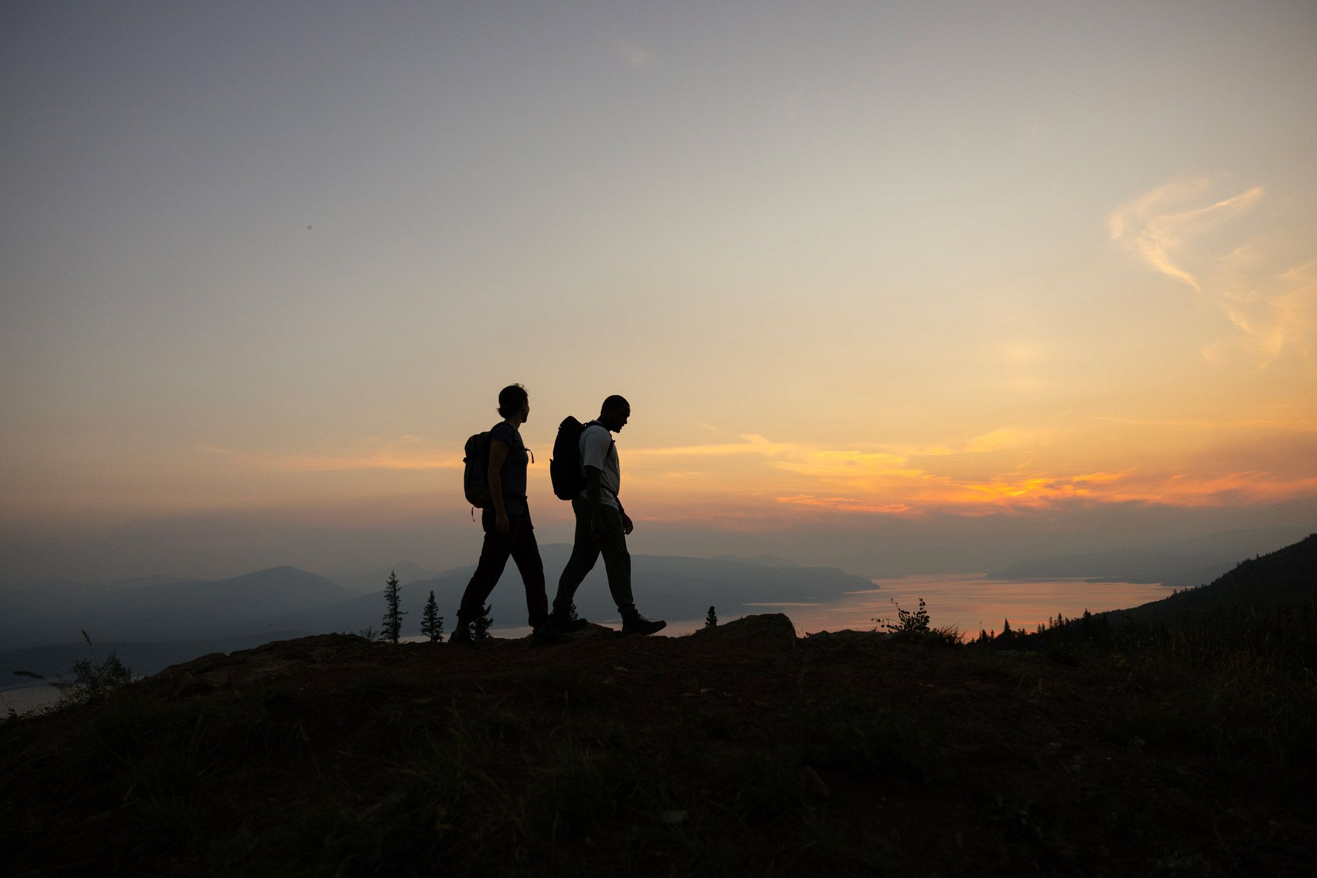 Two hikers traverse a hilltop in a mountainous setting, with a setting sun in the background