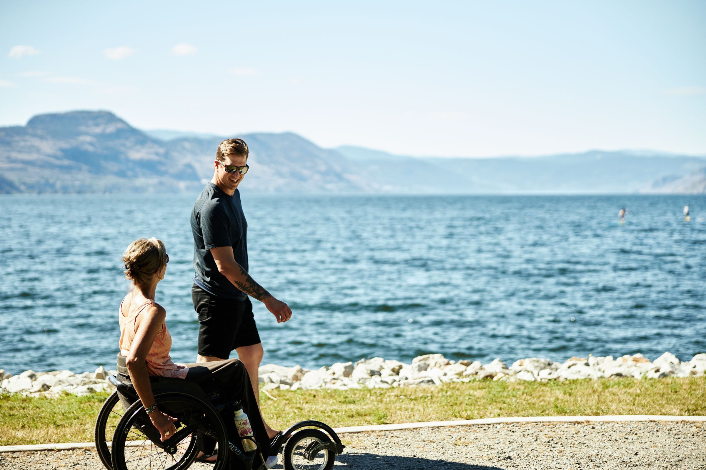A person walks with someone using a wheelchair on a waterside path
