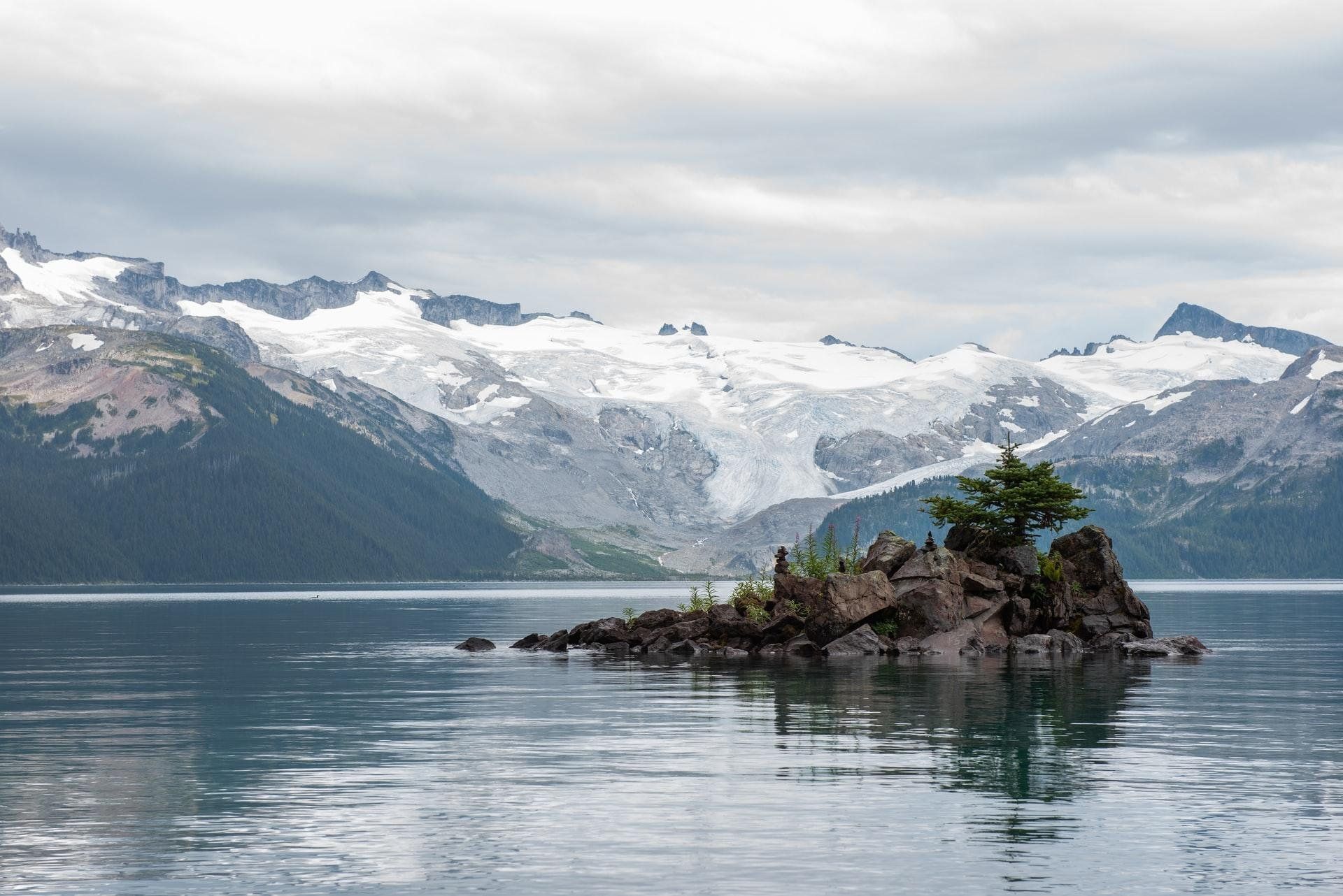 Small rocky island in a calm lake with snowy mountains in the background.