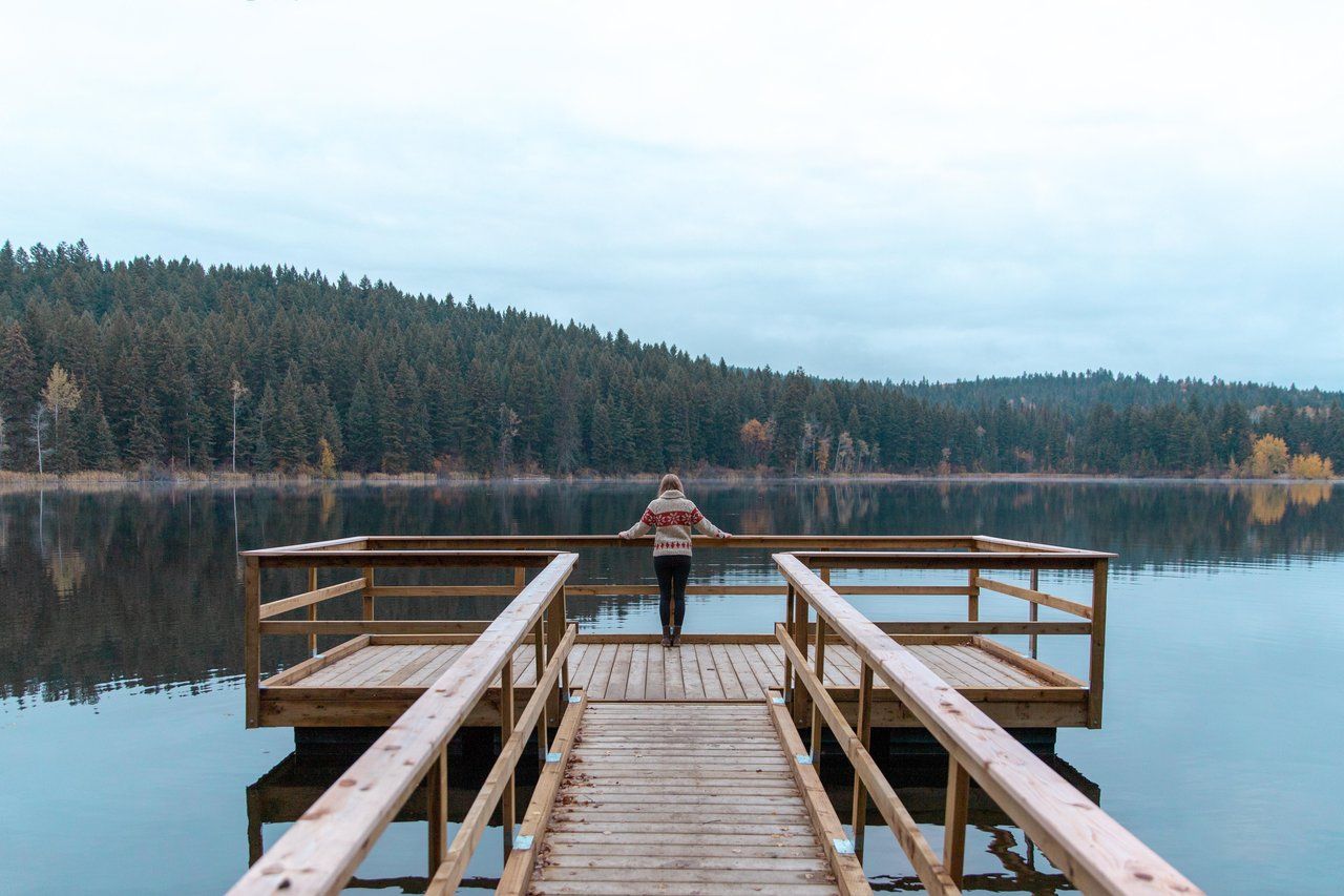 A woman at the end of a wooden dock looking out over a calm, forest-lined lake.