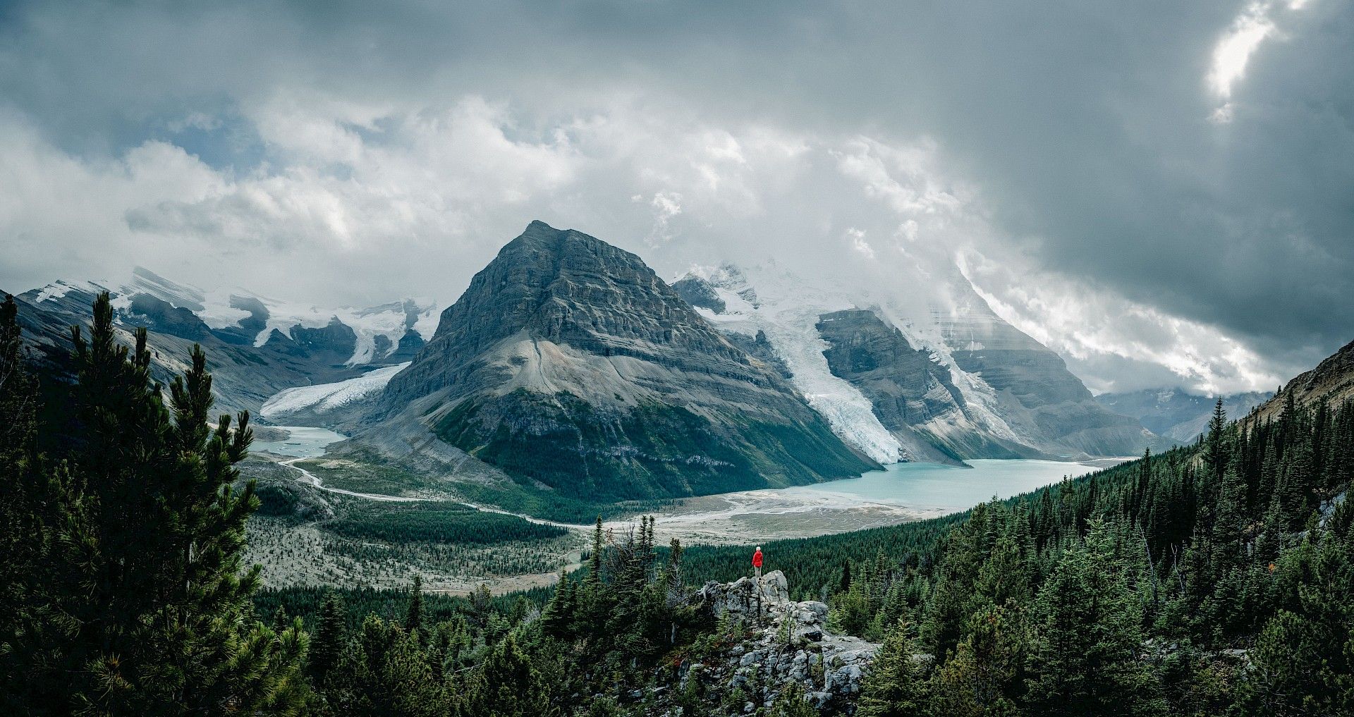 A lone person stands on a rock amongst nature, with large glacial mountains in the surrounding.