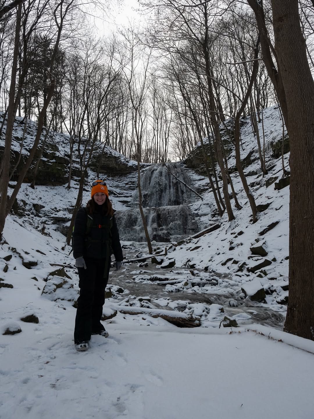 Person on hike poses for picture in front of snowy landscape and frozen waterfall