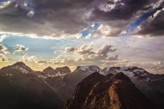 Two people stand upon a mountaintop with a surrounding mountainous landscape and swirling clouds