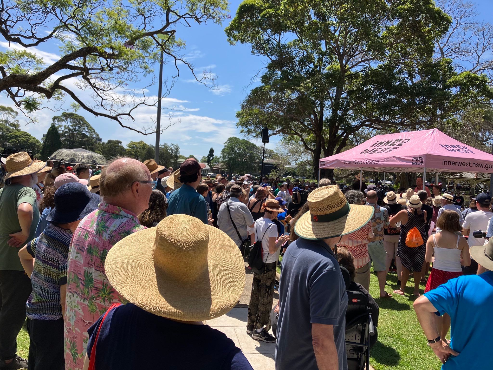 A photograph of a large group of people gathering in a sunny park, everyone is facing towards a tent with a pink roof, listening to a speech. Trees and blue sky is visible in the background.