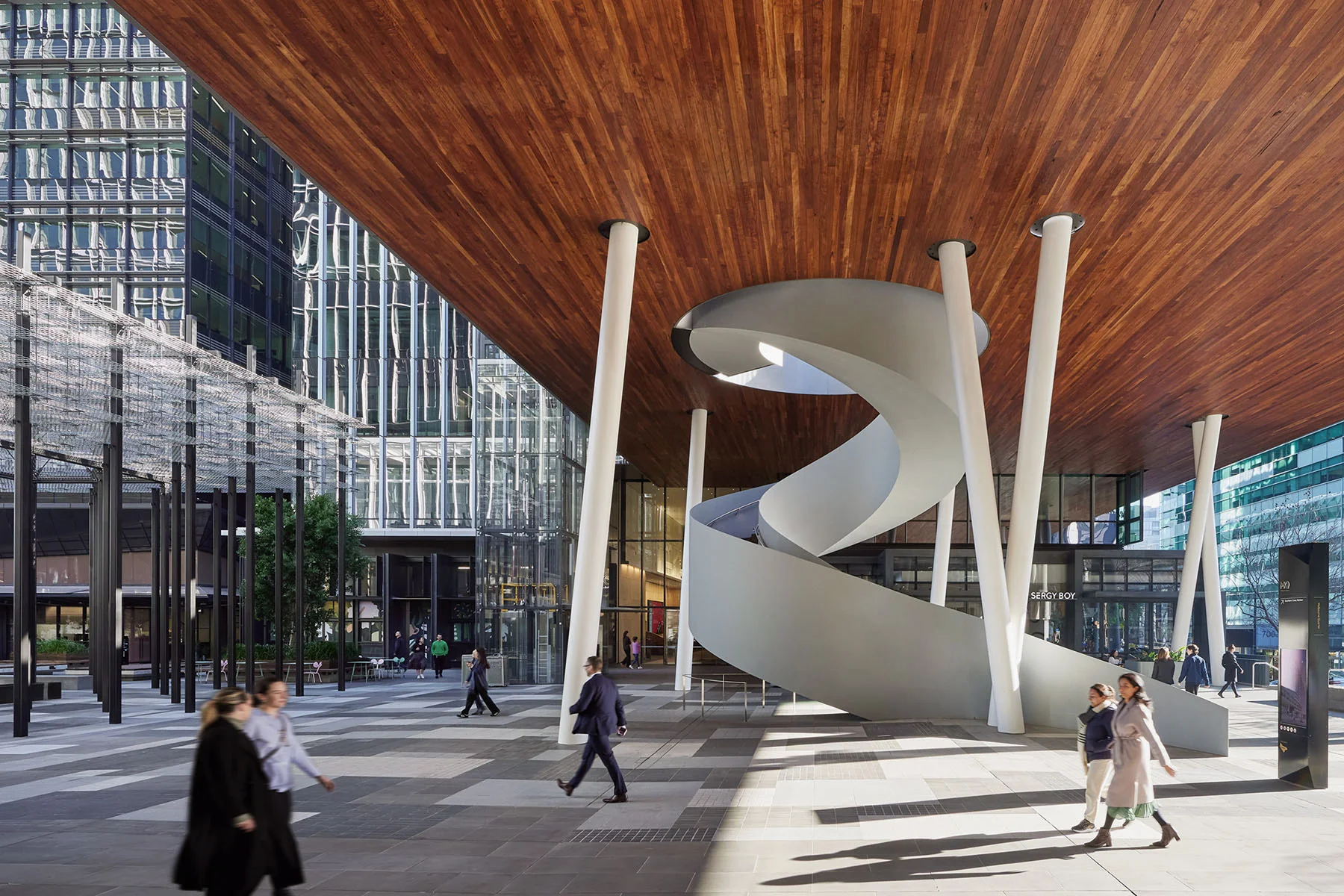 A photo people walking through an urban, outdoor, public space with a significant spiral staircase extending to to a high timber ceiling.