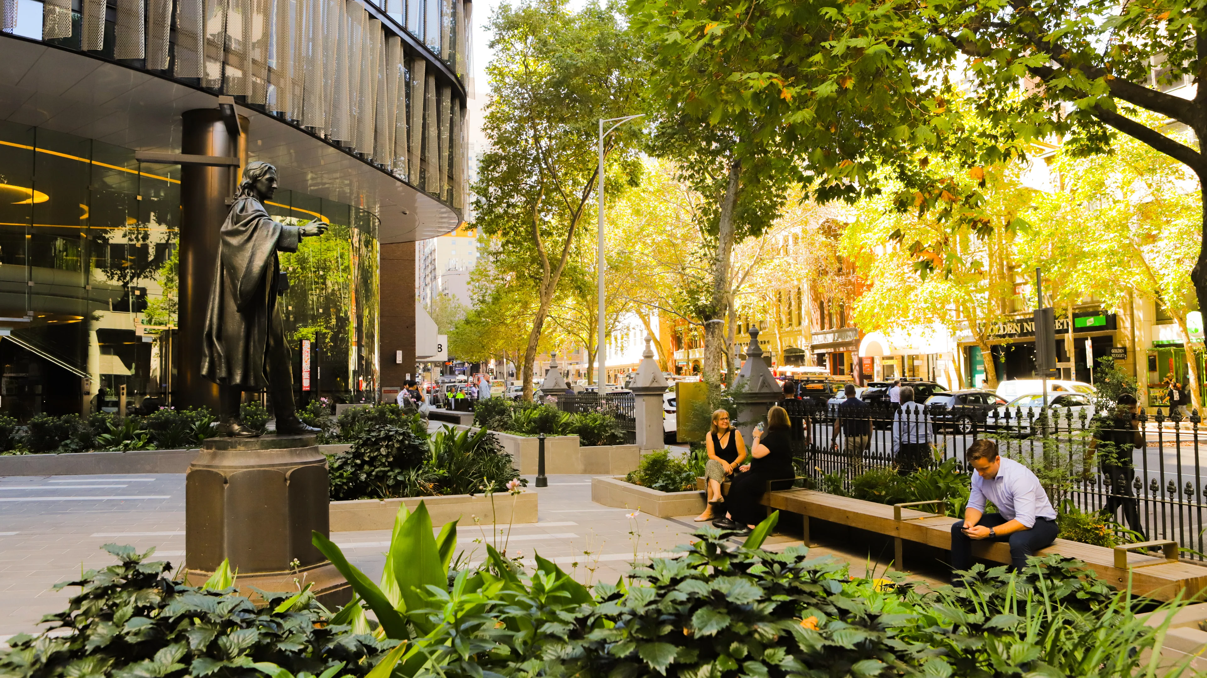A photograph of a few people sitting in an urban forecourt with planting in the foreground, tree canopy above, and a contemporary commercial building to the left.