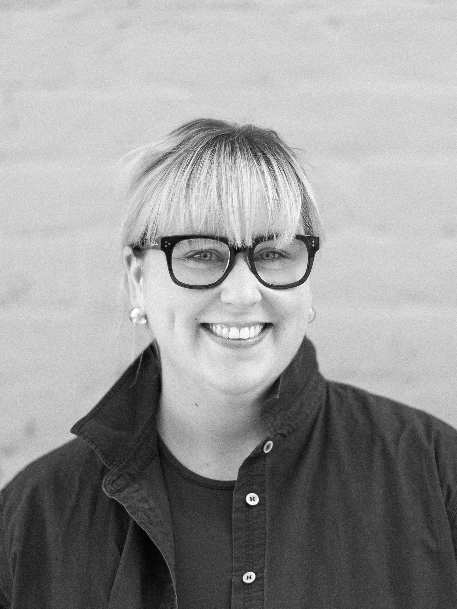 A black and white headshot of a smiling person taken in front of a white brick wall