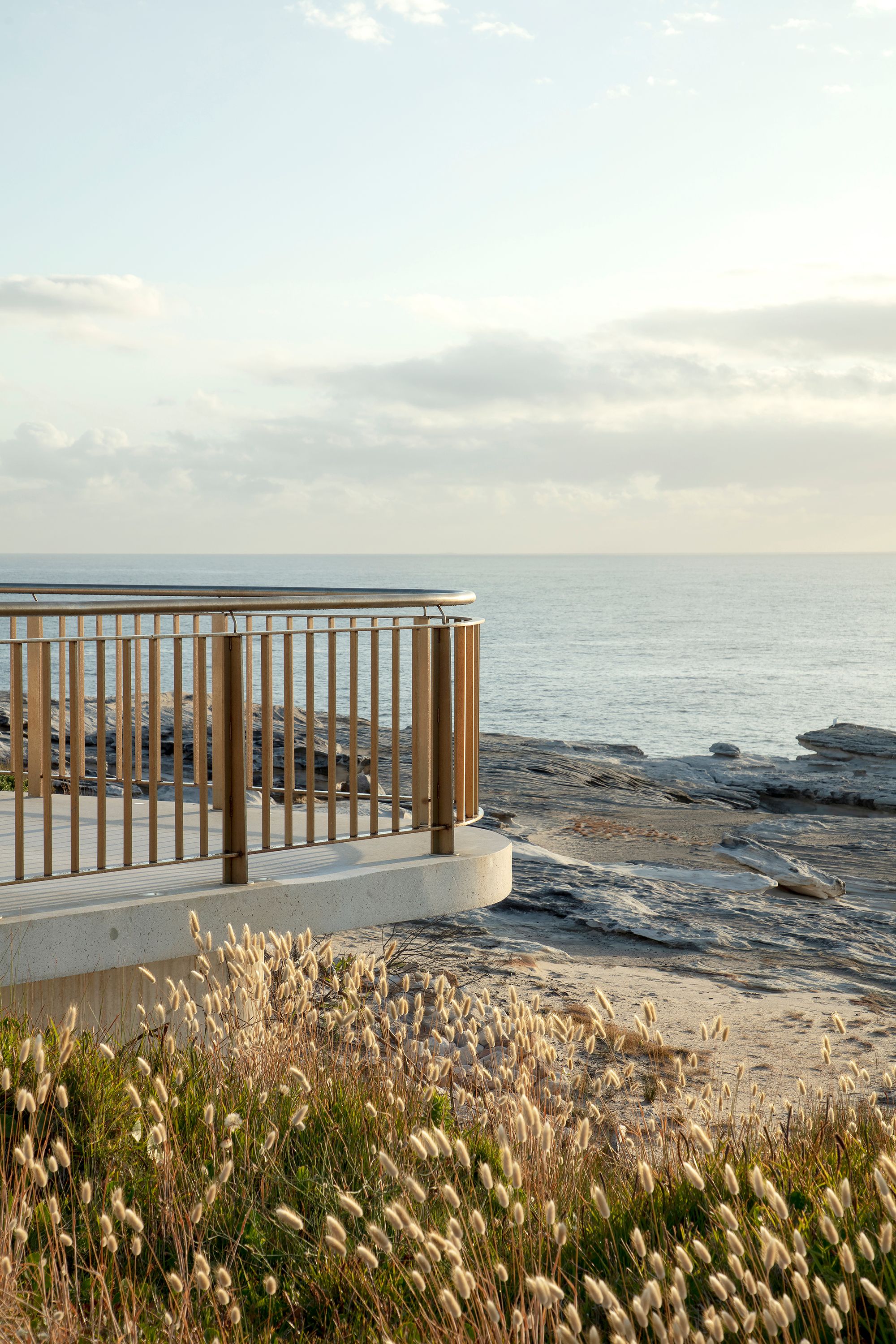 A photograph of the end of curved viewing platform, surrounded by native grasses and rocky cliffs, with a pastel sea and soft cloudy sky in the background.