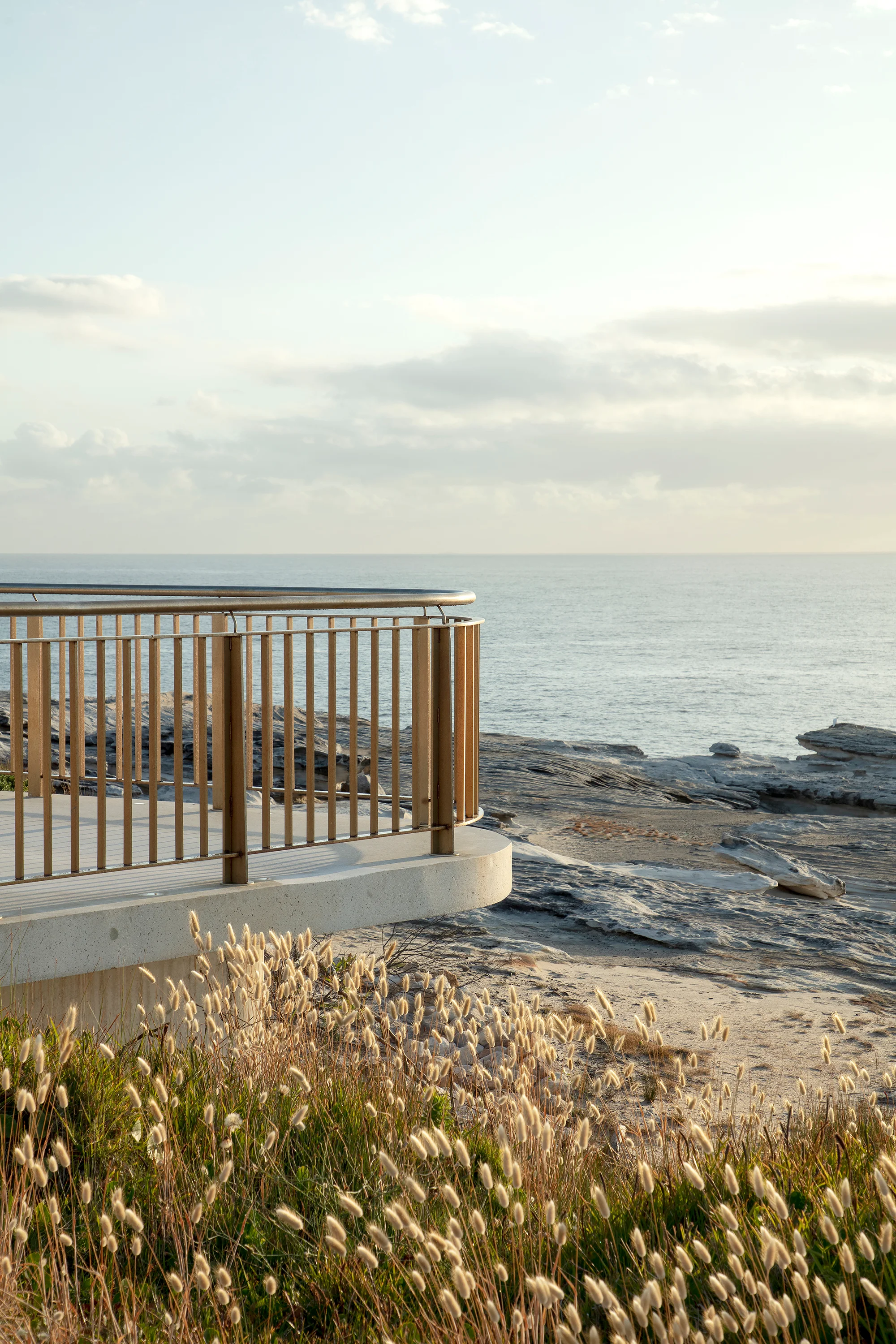 A photograph of the end of curved viewing platform, surrounded by native grasses and rocky cliffs, with a pastel sea and soft cloudy sky in the background.