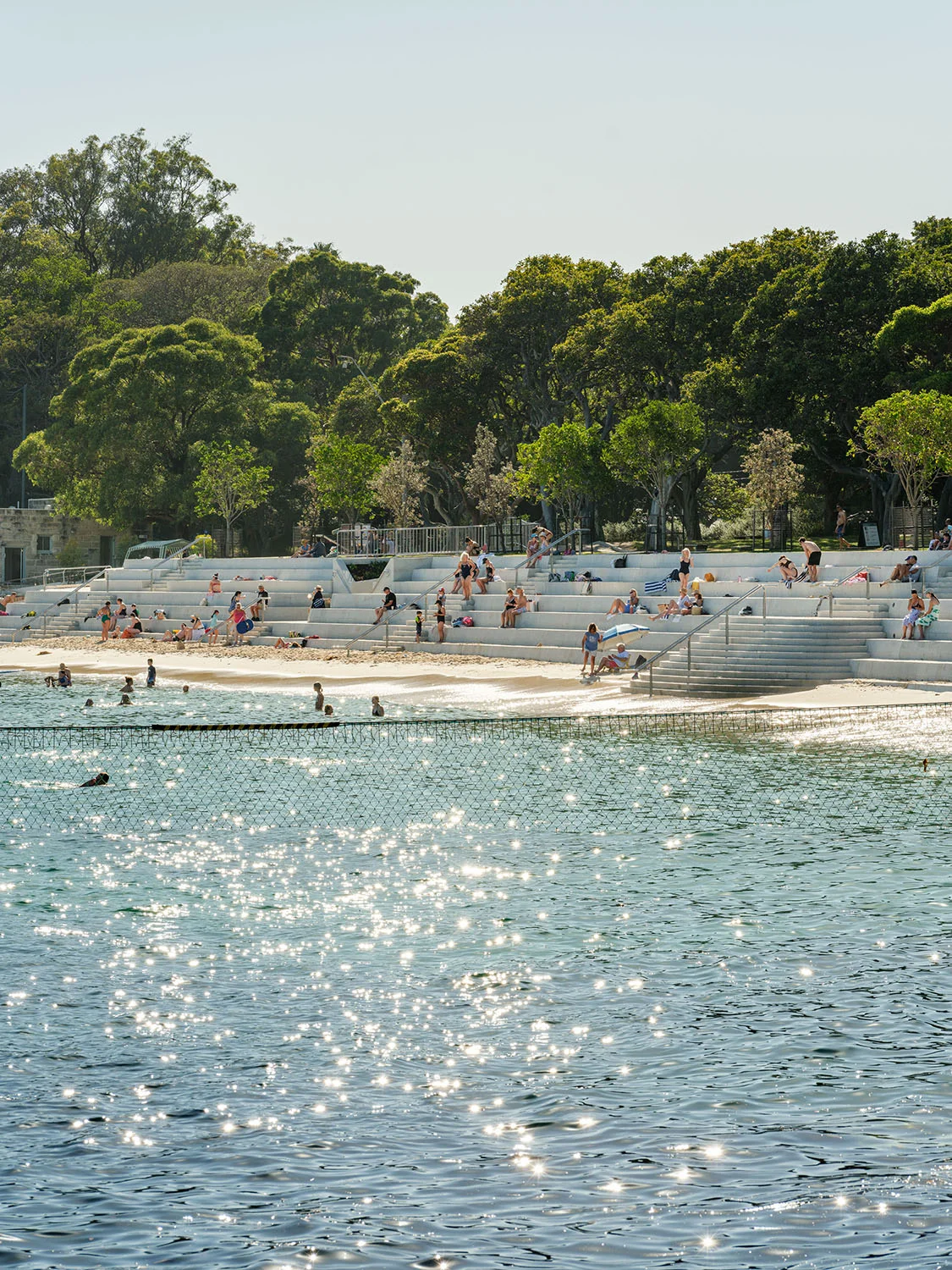 A photograph of a tiered seawall taken across sunny, sparkling water. There are people sitting, standing, and swimming.