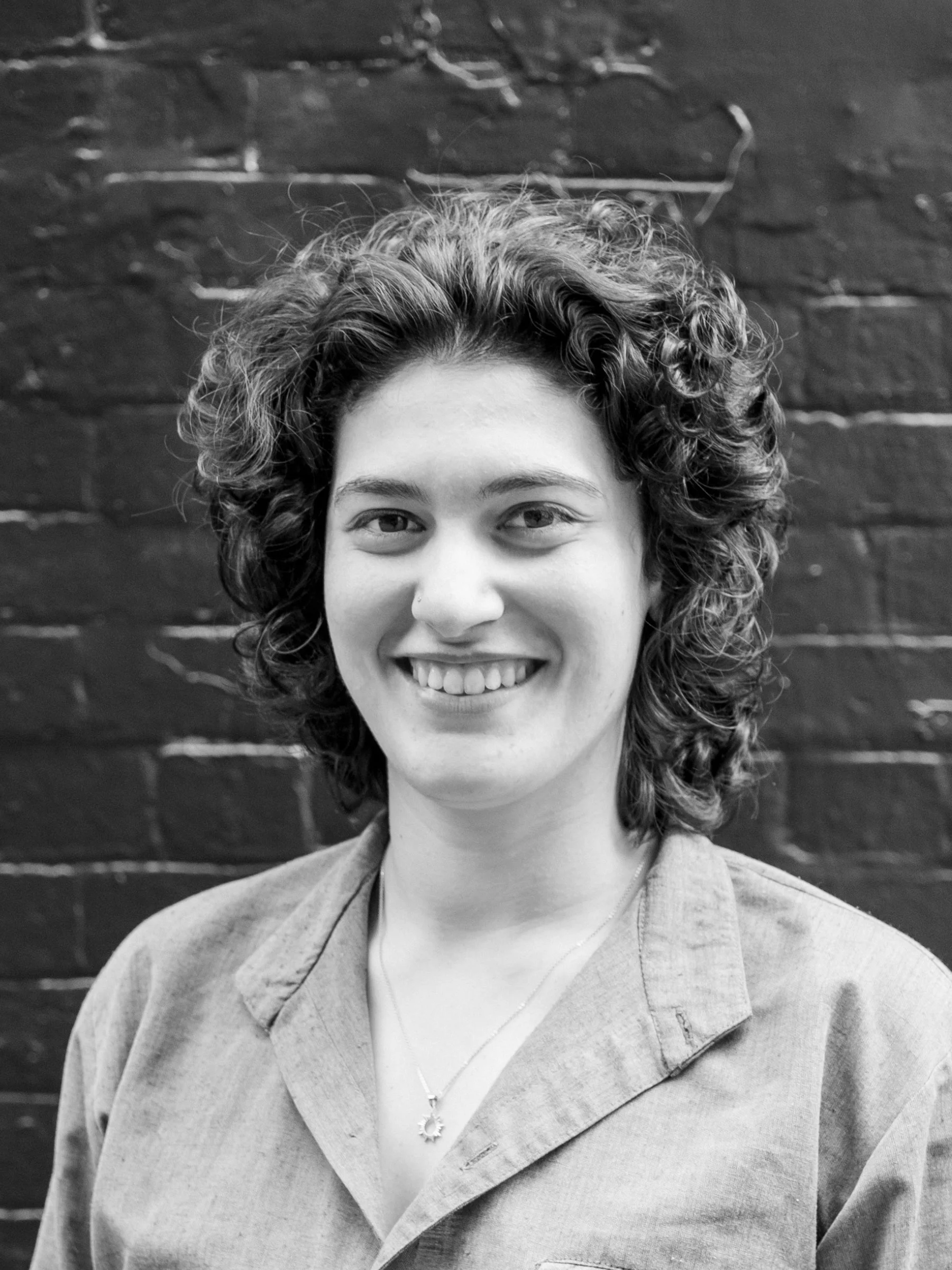 A black and white headshot of a person with curly hair in front of a brick wall painted black