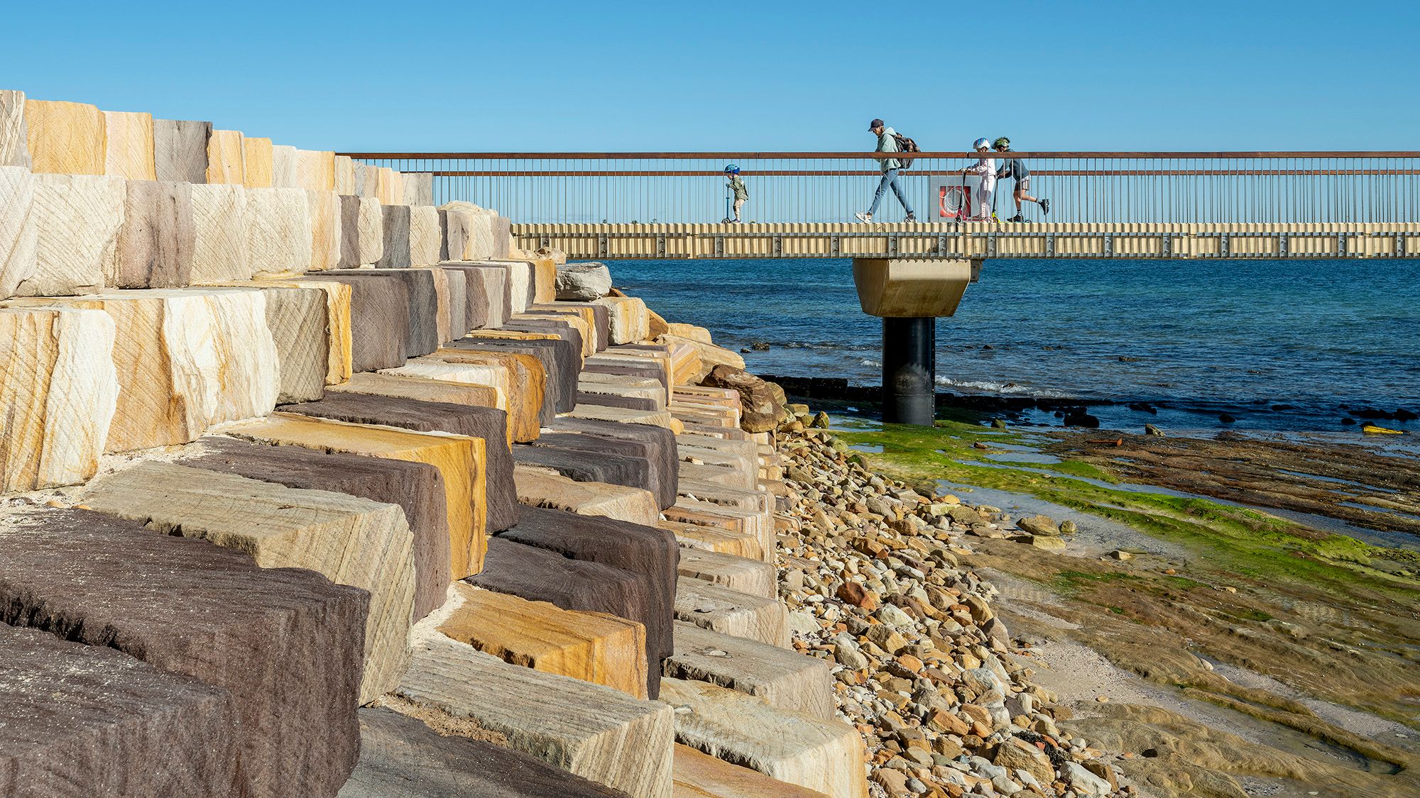 People walk across a bridge with a revetment wall shown in the foreground.