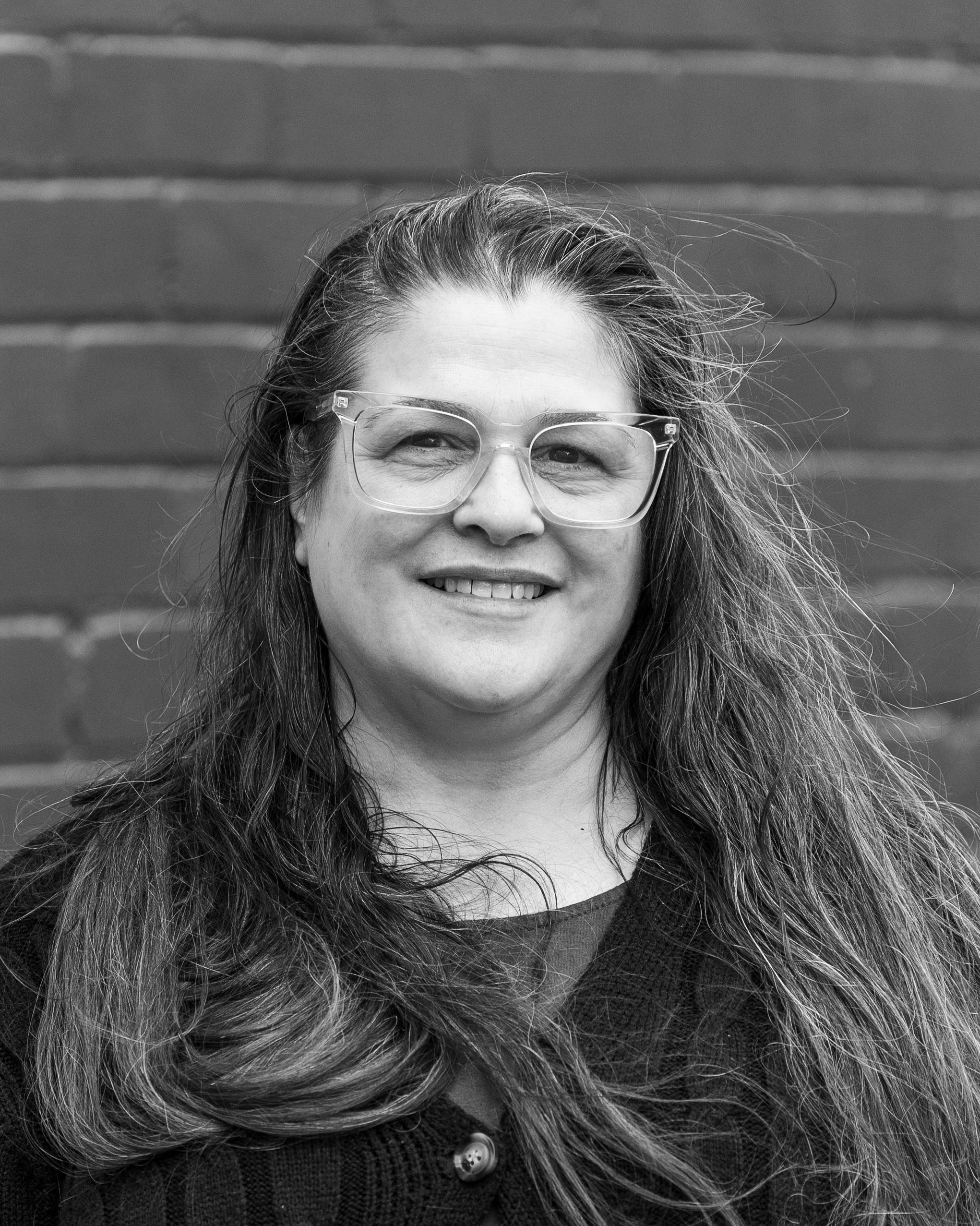 A black and white headshot of a smiling person in front of a brick wall.