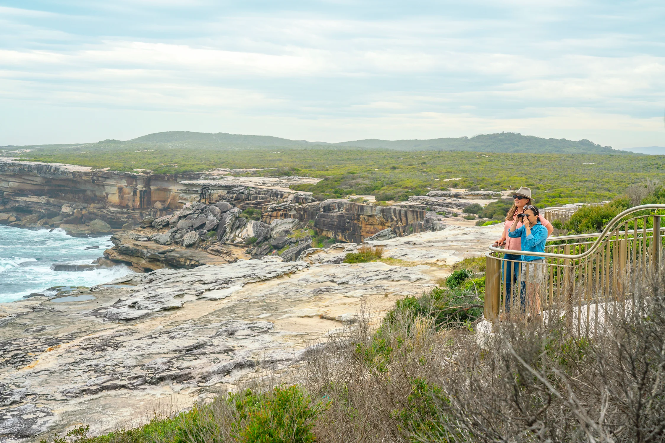 A photograph of two people standing on a viewing platform overlooking a dramatic sandstone coastline. One is wearing a hat, the other is looking through binoculars. There is vegetation in the foreground and into the distance.