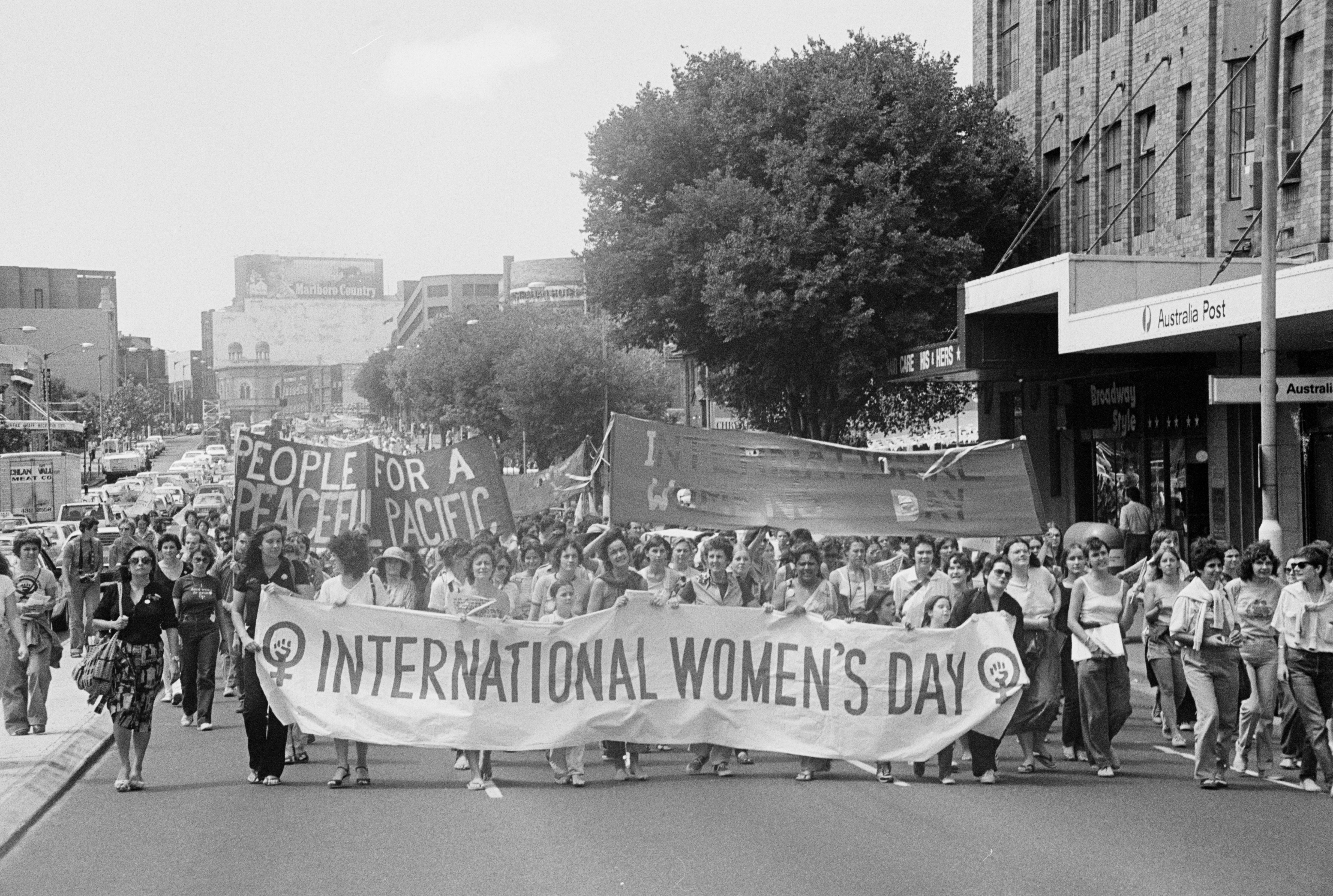 A black and white photograph (scanned from film) of an International Women's Day March in Sydney in the 1980's. People are marching towards the camera, a banner stretches across the street reading 'International Women's Day'. The crowd stretches down the street, disappearing into the background of the photo.