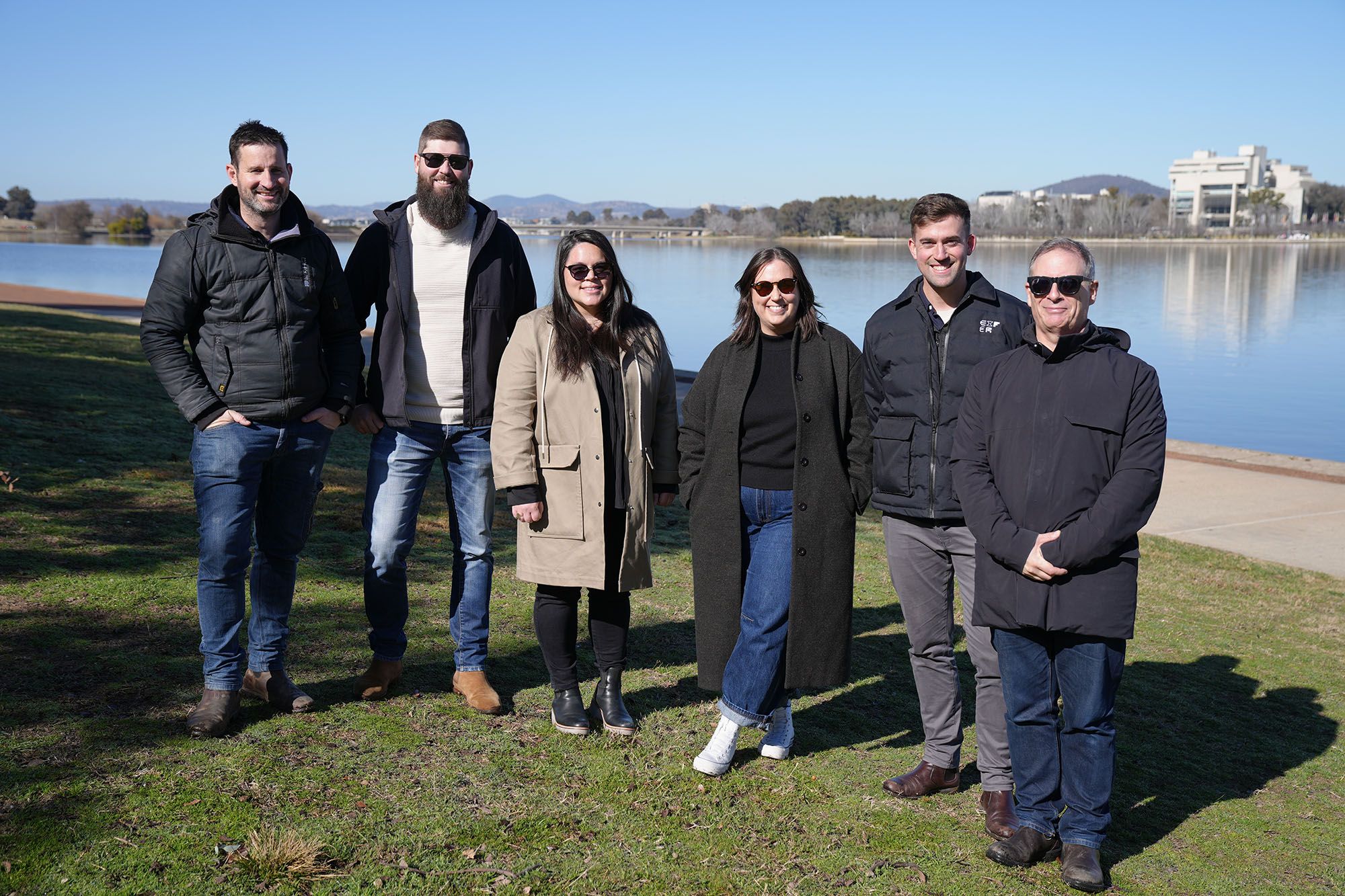 A photo of six people standing in a row on a patch of grass smiling for the camera. There is a lake behind, and they are all wearing warm jackets. Four a wearing sunglasses.